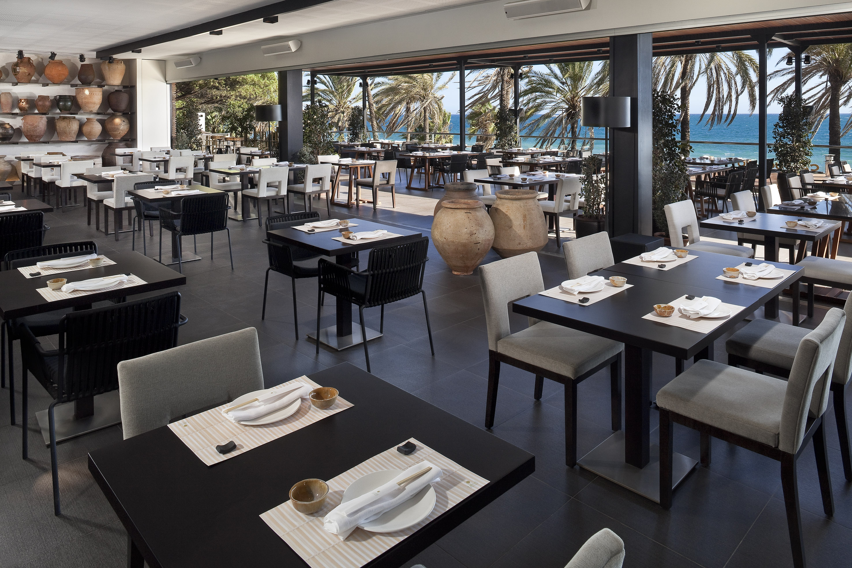 Spacious oceanfront restaurant with palm trees, blue sea, and modern dining tables.