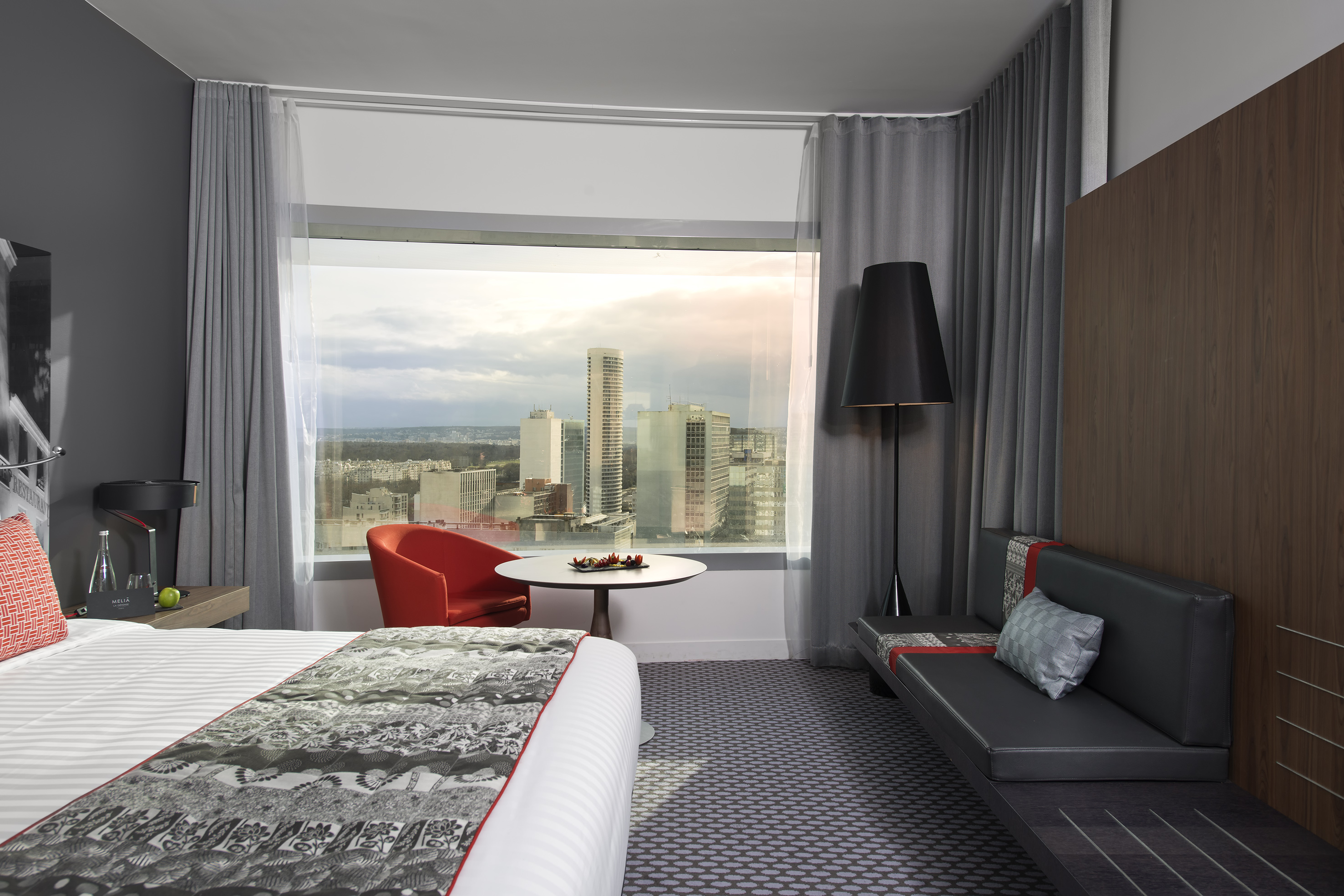 Modern hotel room with city skyline view, bed, red armchair, and seating.