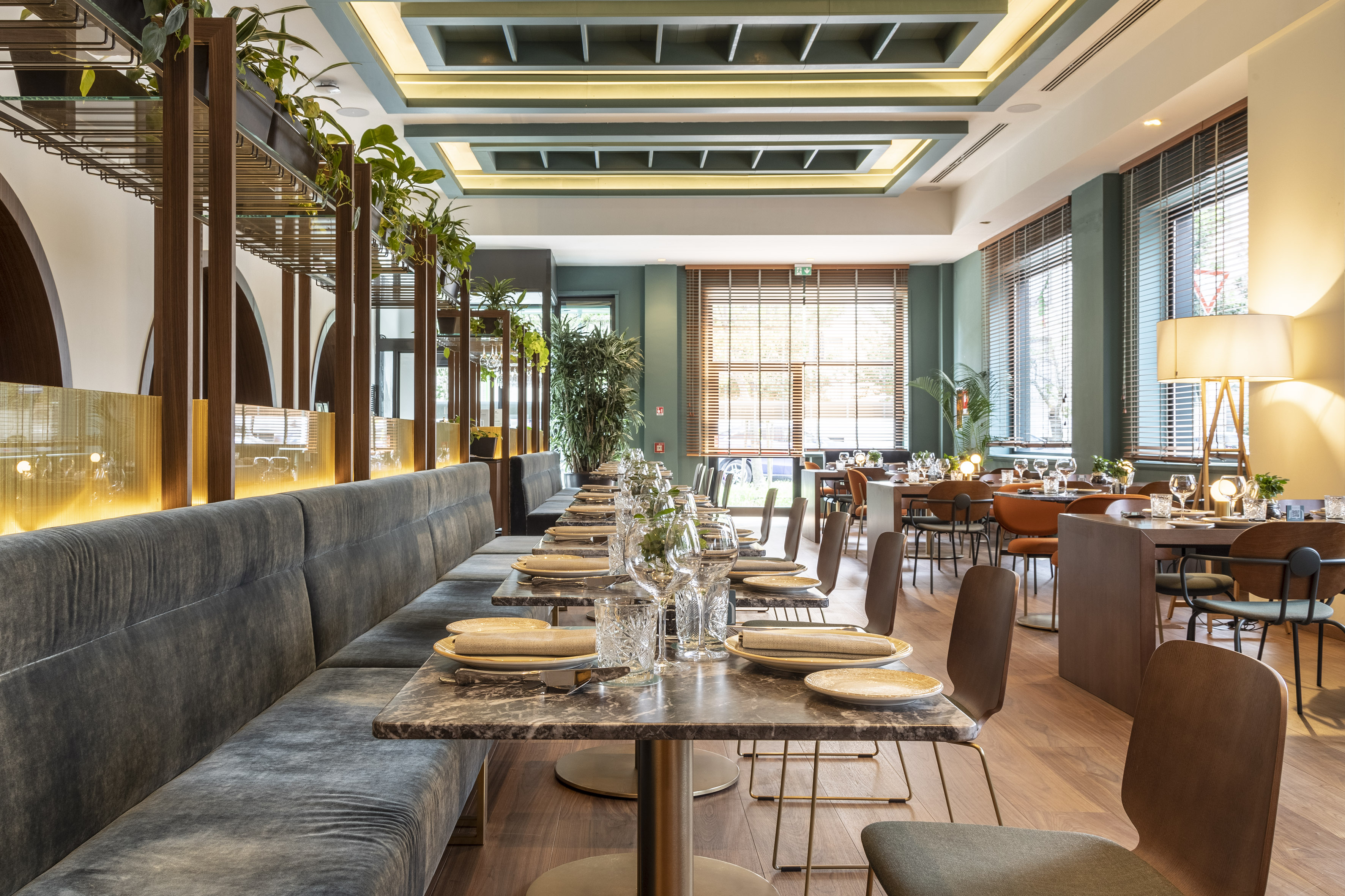 Modern restaurant interior with velvet banquette, marble tables, and green plants.