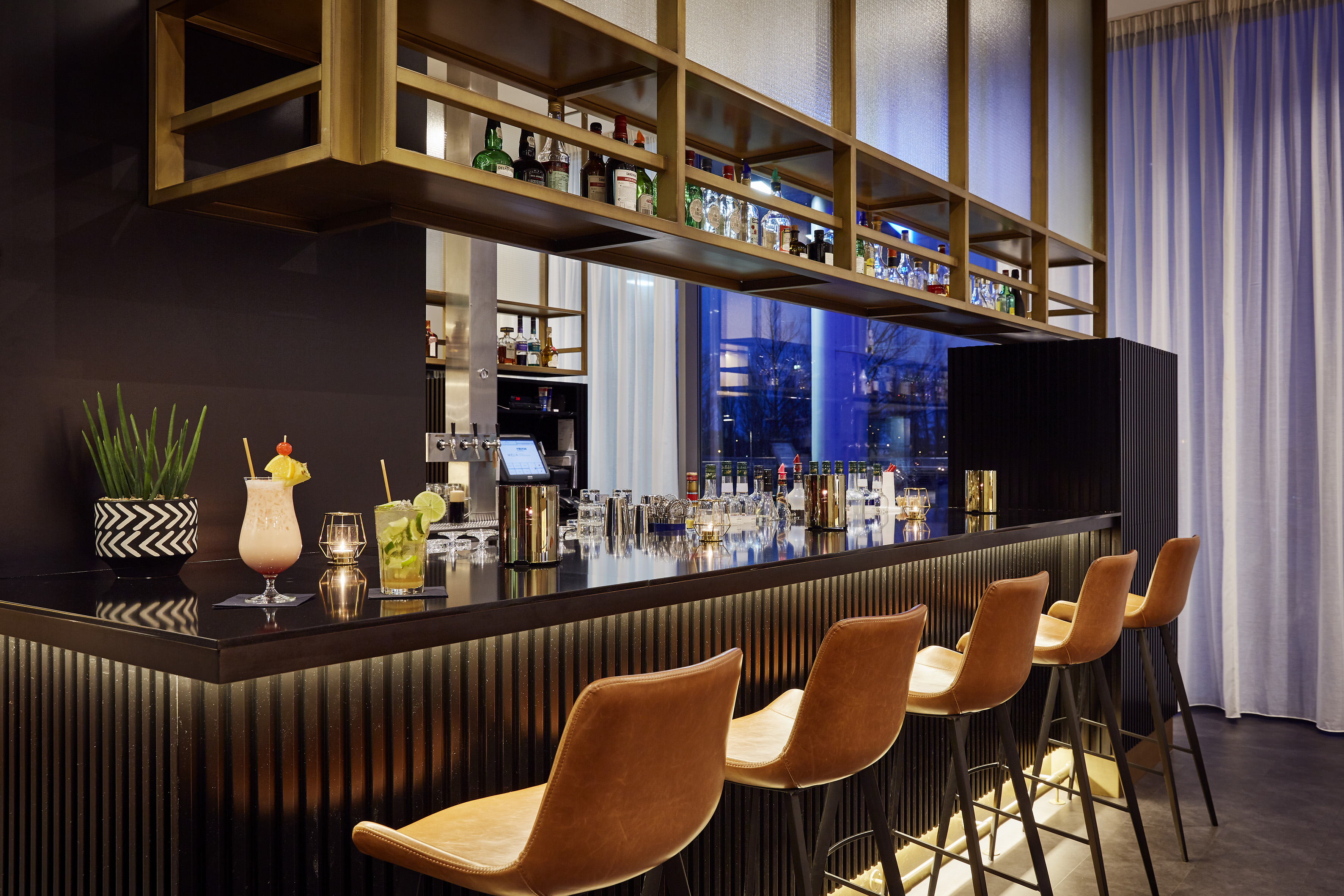Sleek hotel bar with cocktails, brown leather stools, and backlit liquor display.
