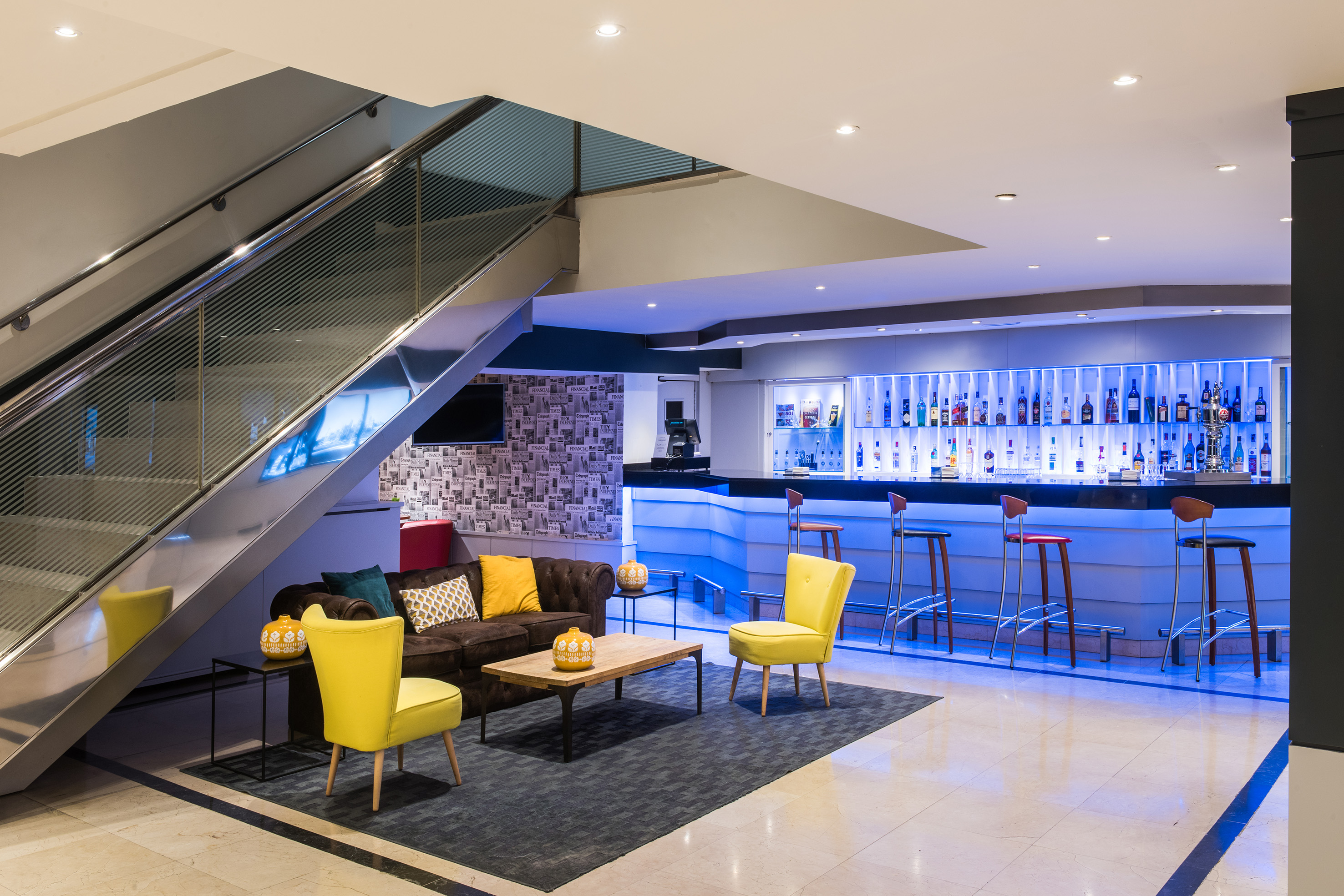 Modern hotel lounge with blue-lit bar, brown sofa, yellow chairs, and sleek staircase.