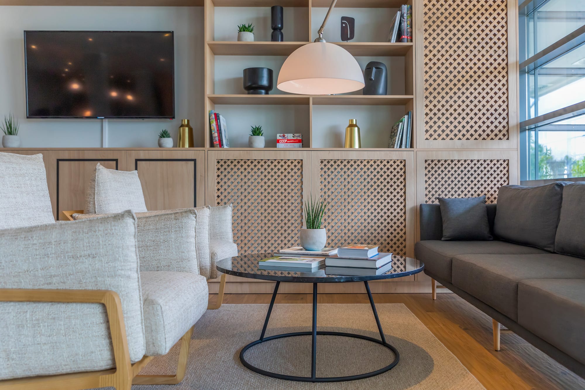 Modern lounge interior with wooden shelving, TV, armchairs, and gray sofa.