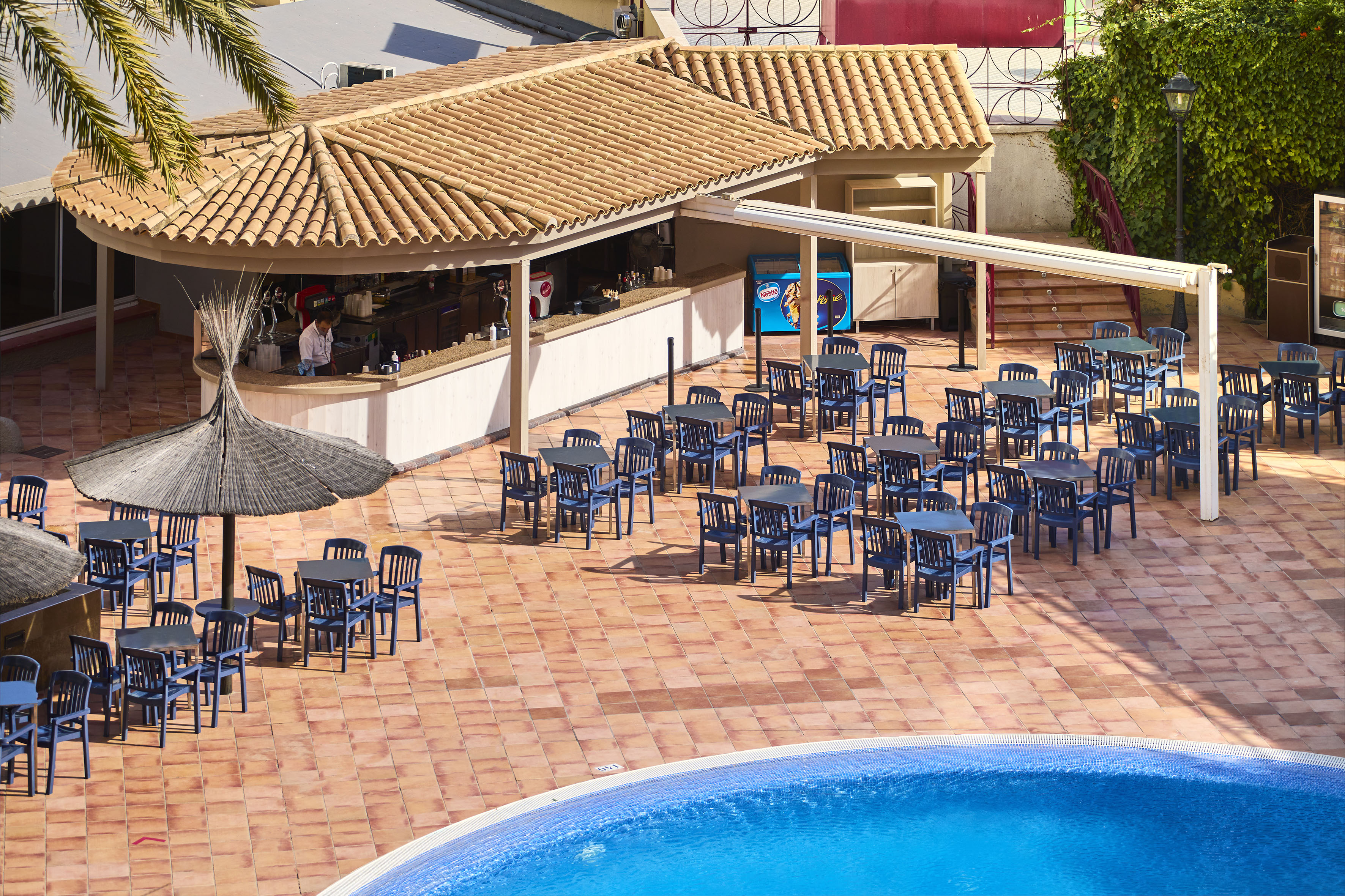 Hotel poolside bar and outdoor dining area with blue tables, chairs, and pool.