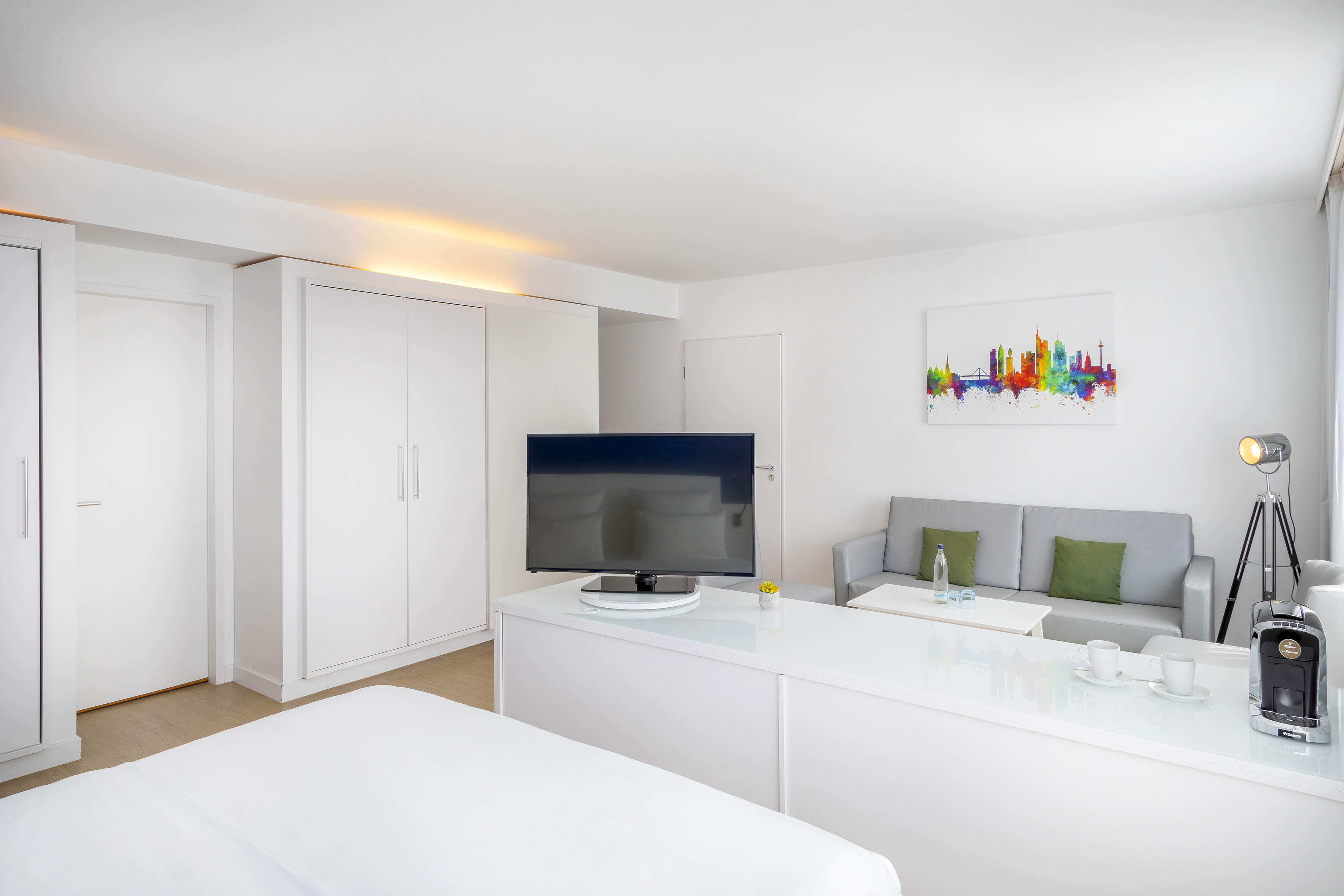 Modern white hotel room with TV, gray sofa, and colorful skyline artwork.