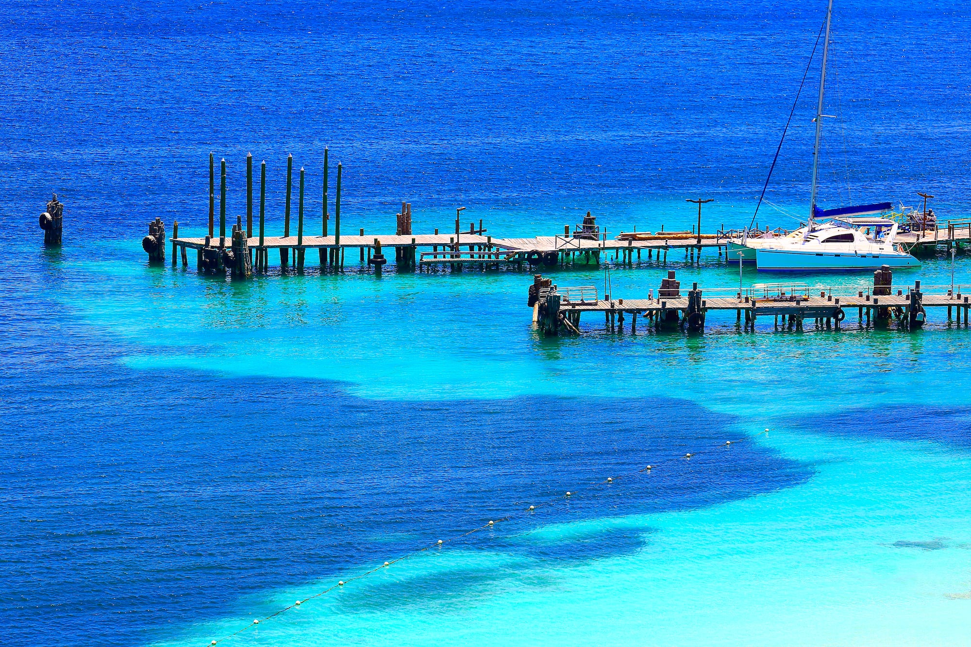 Catamaran sailboat docked at wooden pier in vibrant blue tropical ocean.