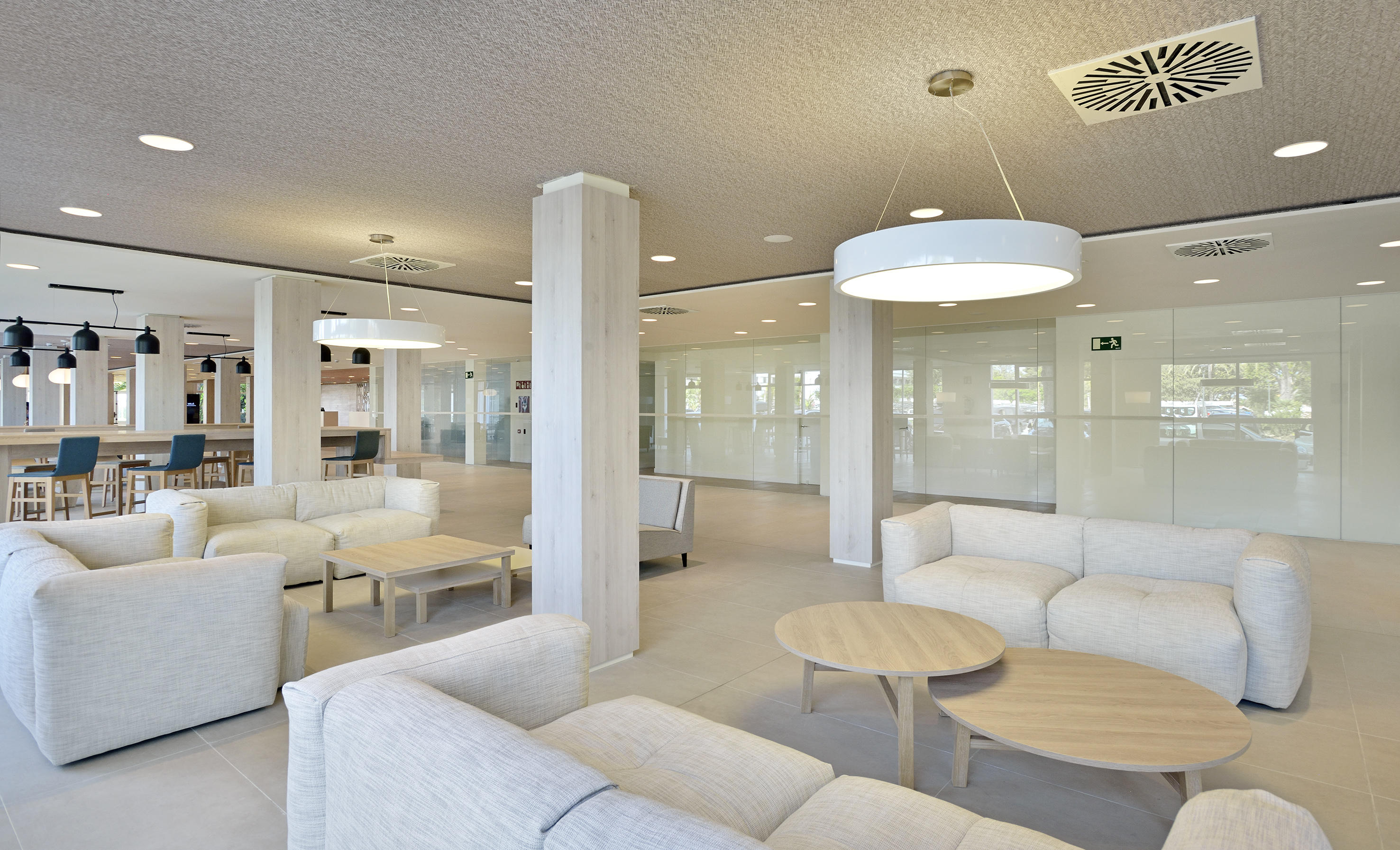 Modern office lobby lounge with light sofas, wooden tables, and glass walls.