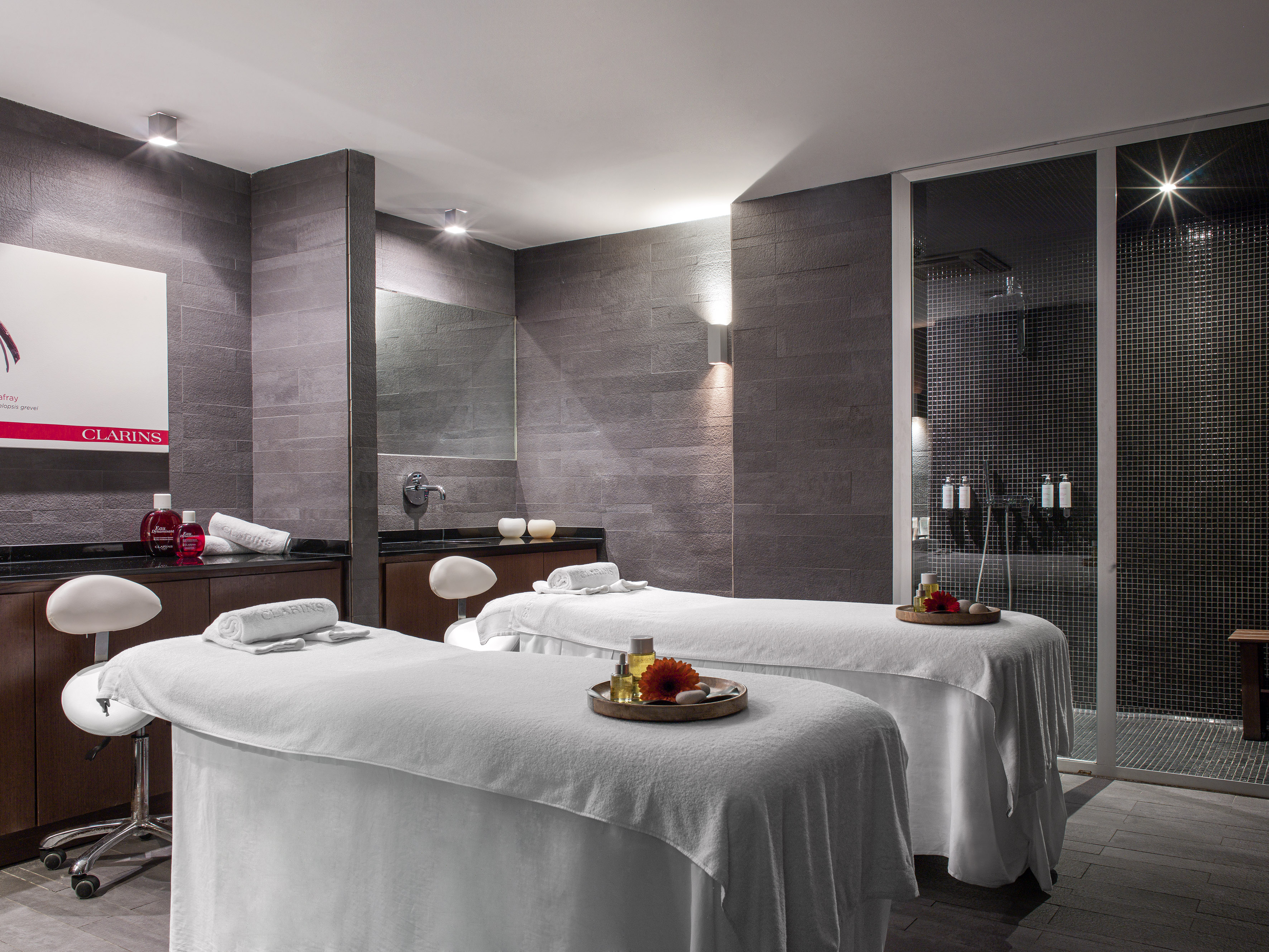Modern spa treatment room with two massage beds, Clarins products, and shower.