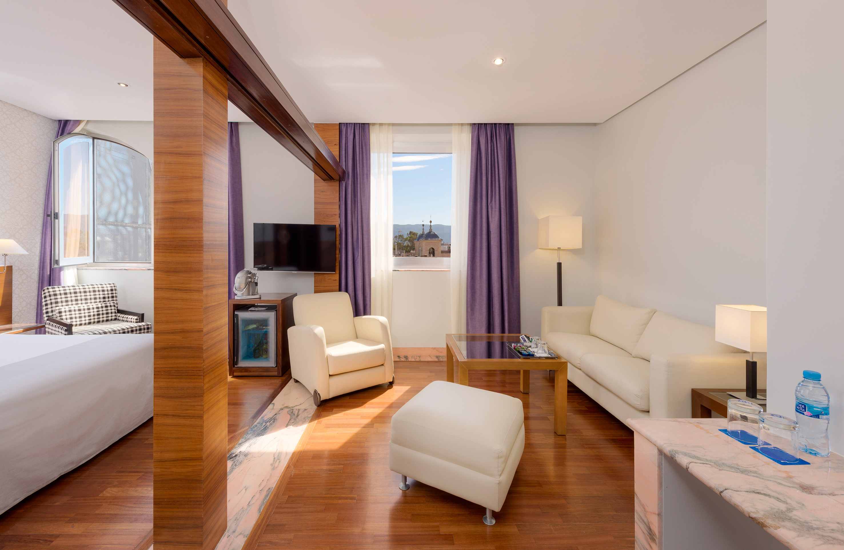 Modern hotel suite living area with white sofa, TV, and city view.