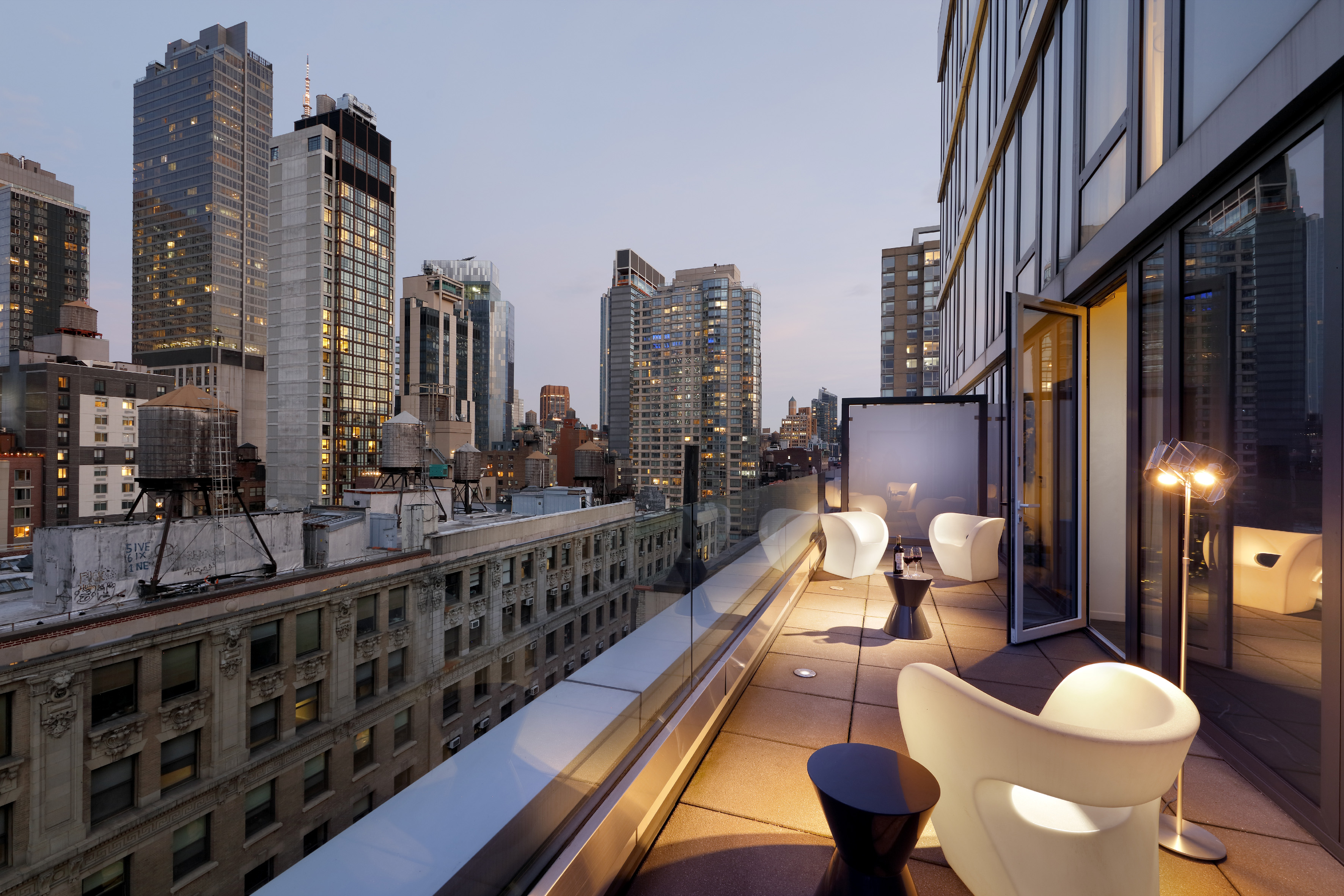 Modern rooftop terrace with white chairs, city skyline, and glass railing at dusk.