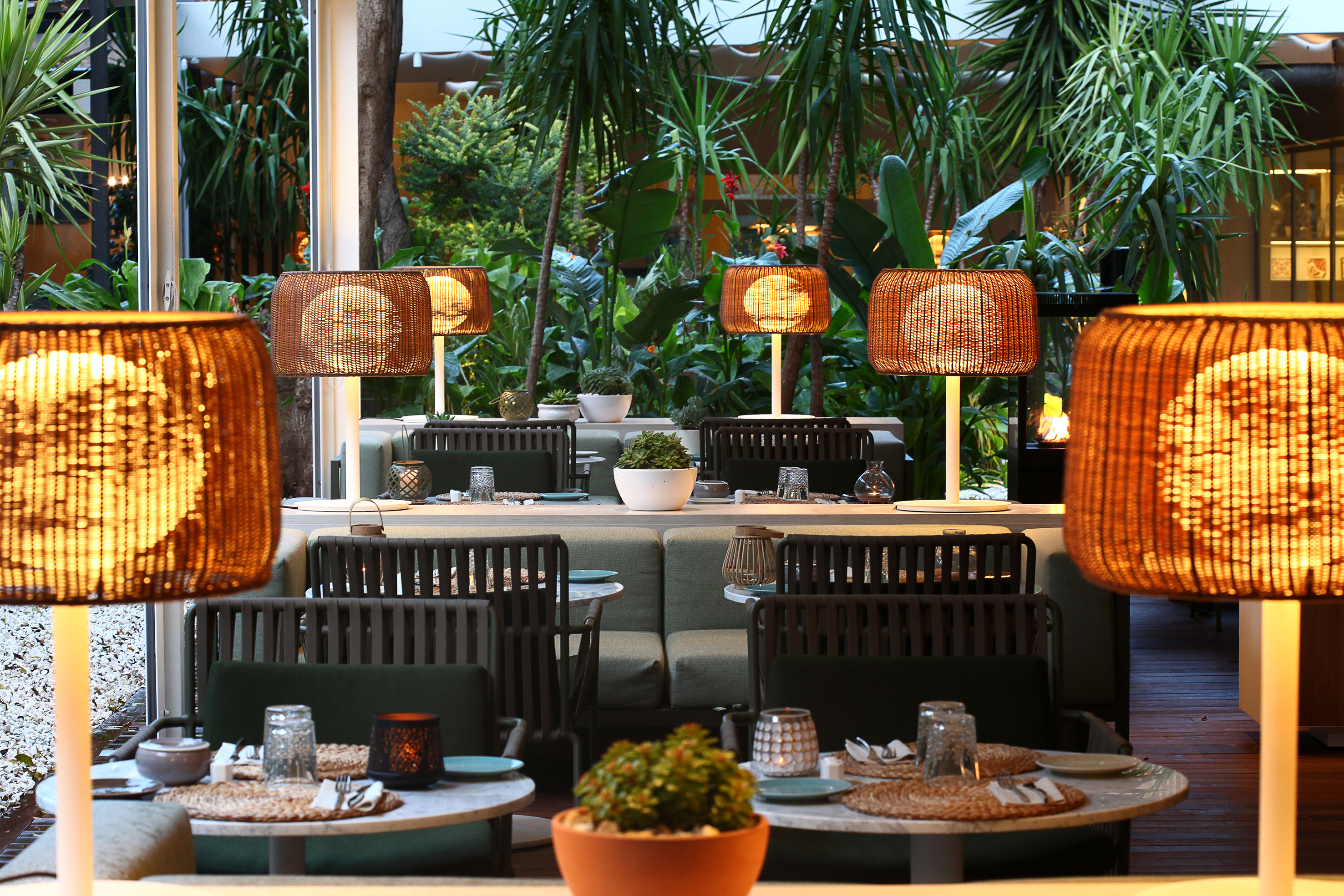 Restaurant patio with rattan table lamps, green banquettes, and tropical garden.