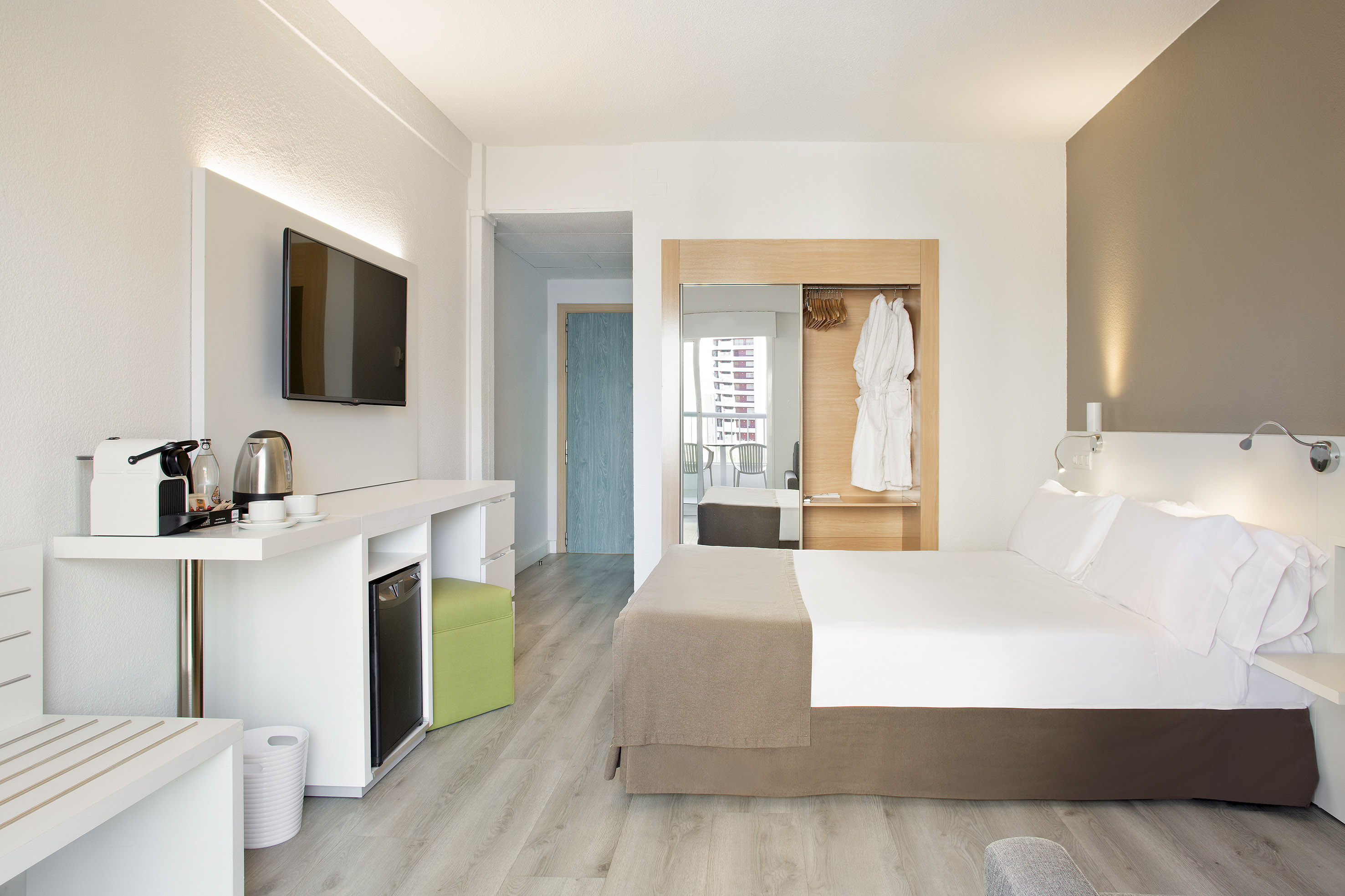Modern hotel room with bed, TV, coffee maker, mini-fridge, and wardrobe.