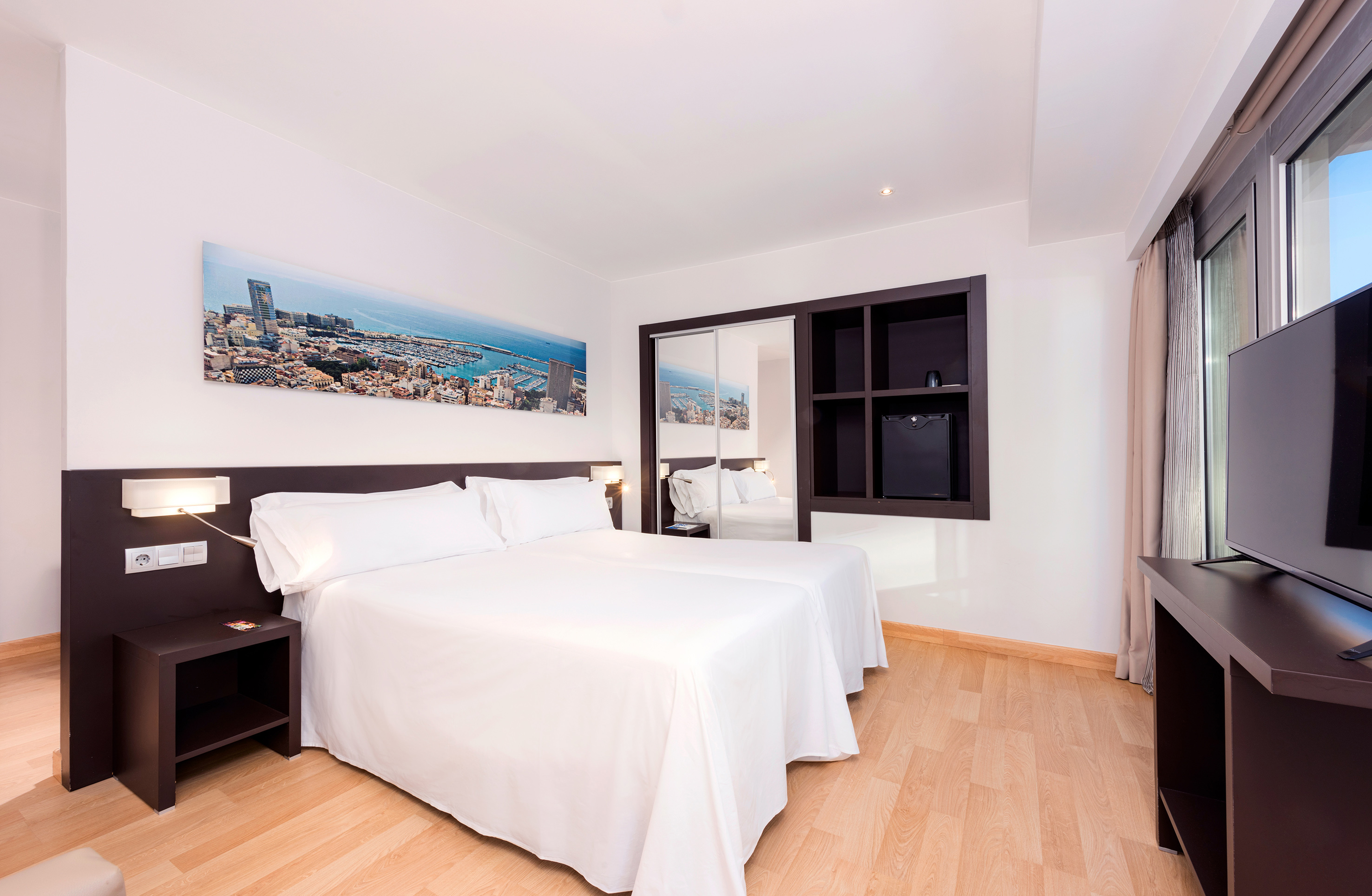 Modern hotel room with twin beds, city art, mirrored closet, and TV.