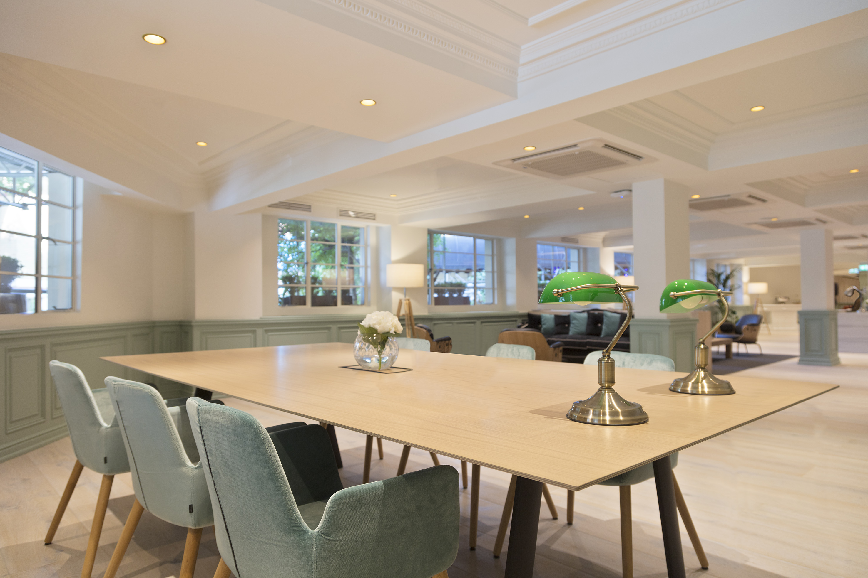 Modern hotel lounge with long wooden table, green chairs, and banker lamps.
