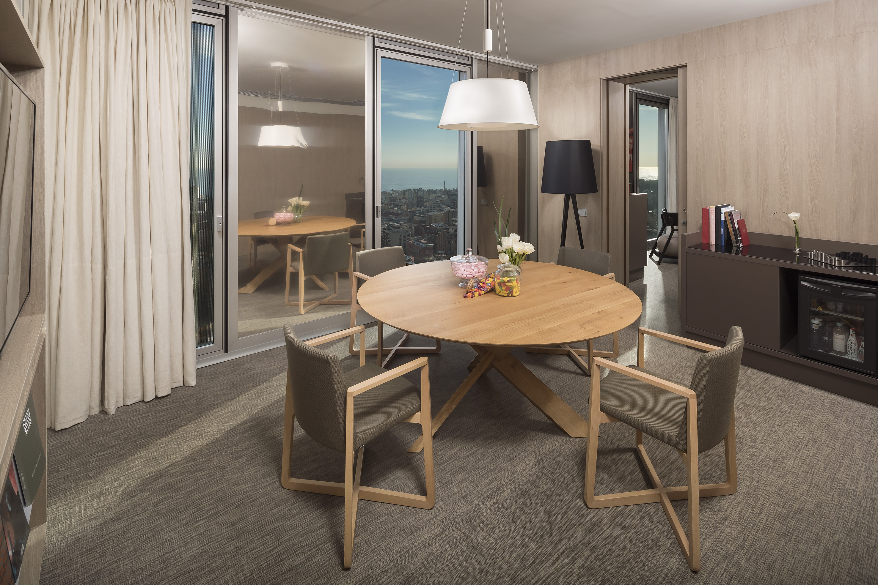 Modern hotel suite dining area with city & sea view, round wooden table.