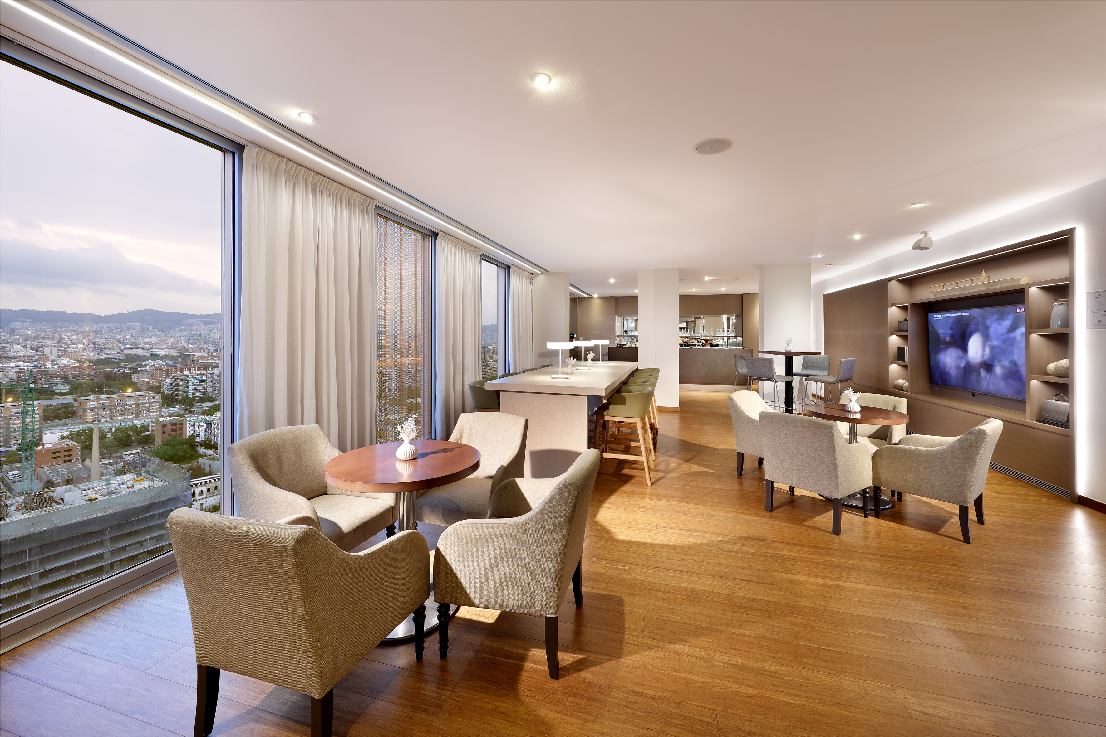Modern lounge with panoramic city view, seating areas, communal table, and TV.