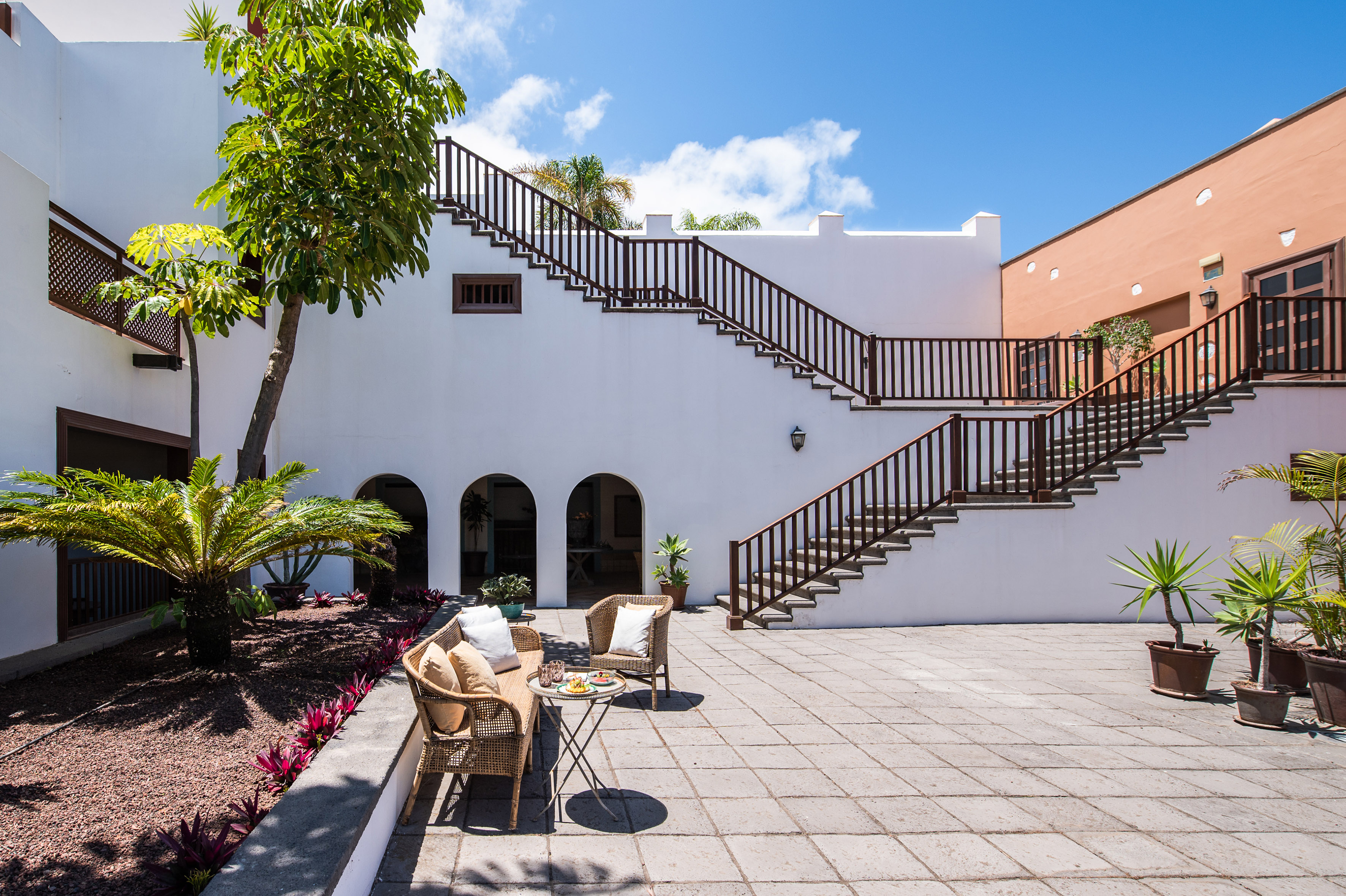 Sunny patio with white architecture, outdoor stairs, seating, and lush plants.