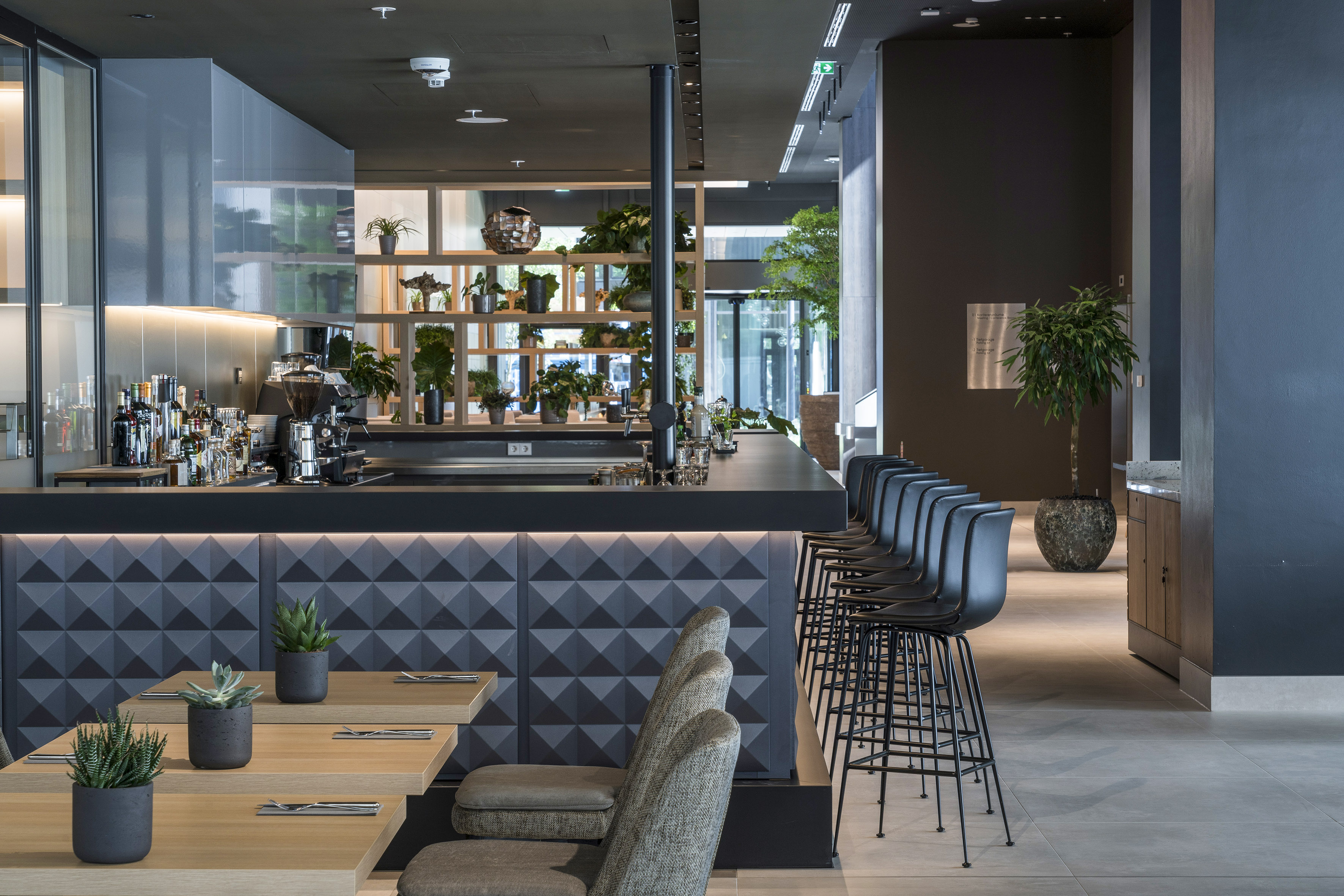 Modern bar interior with geometric facade, black stools, and potted plants.
