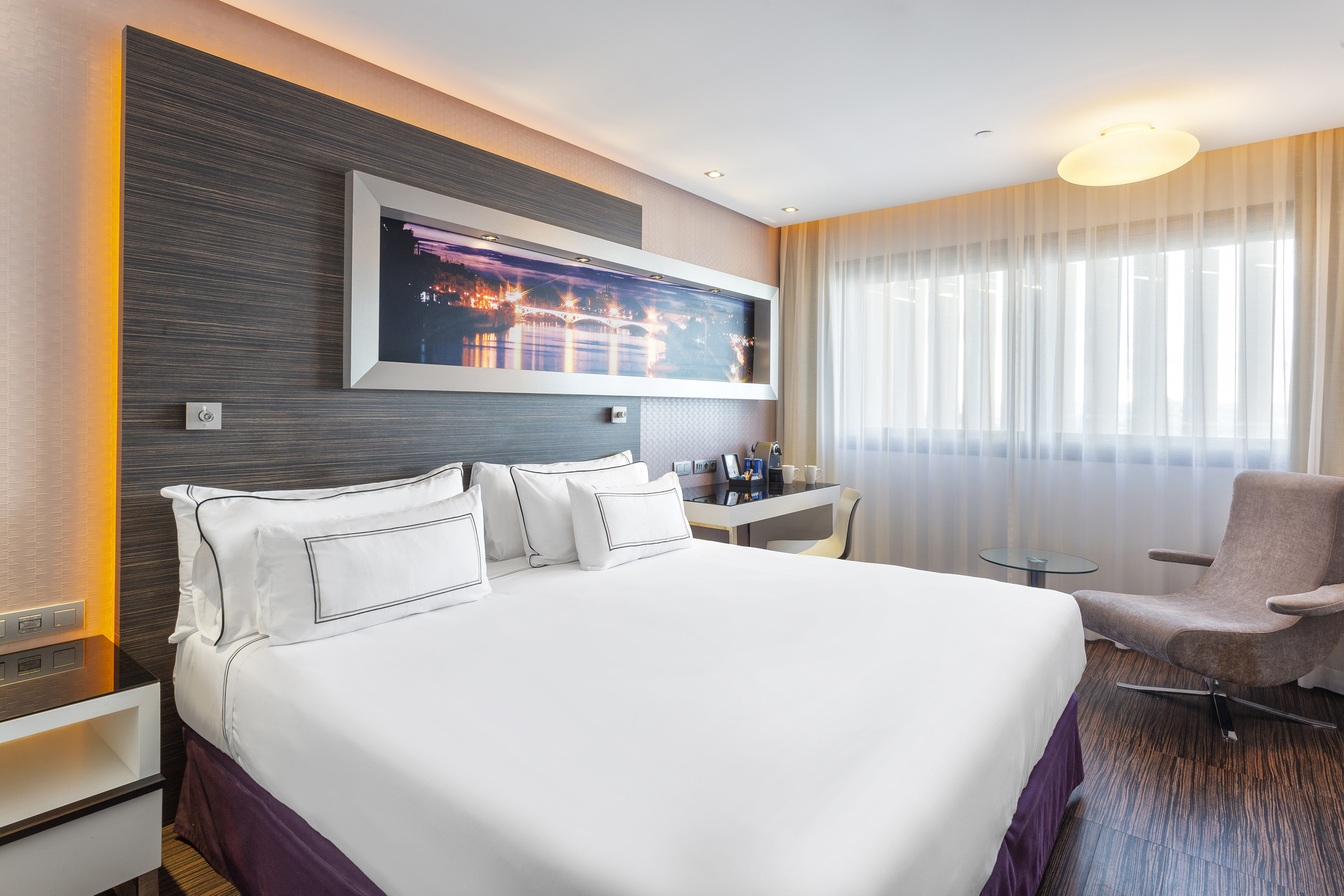 Modern hotel bedroom with king bed, desk, armchair, and city skyline artwork.