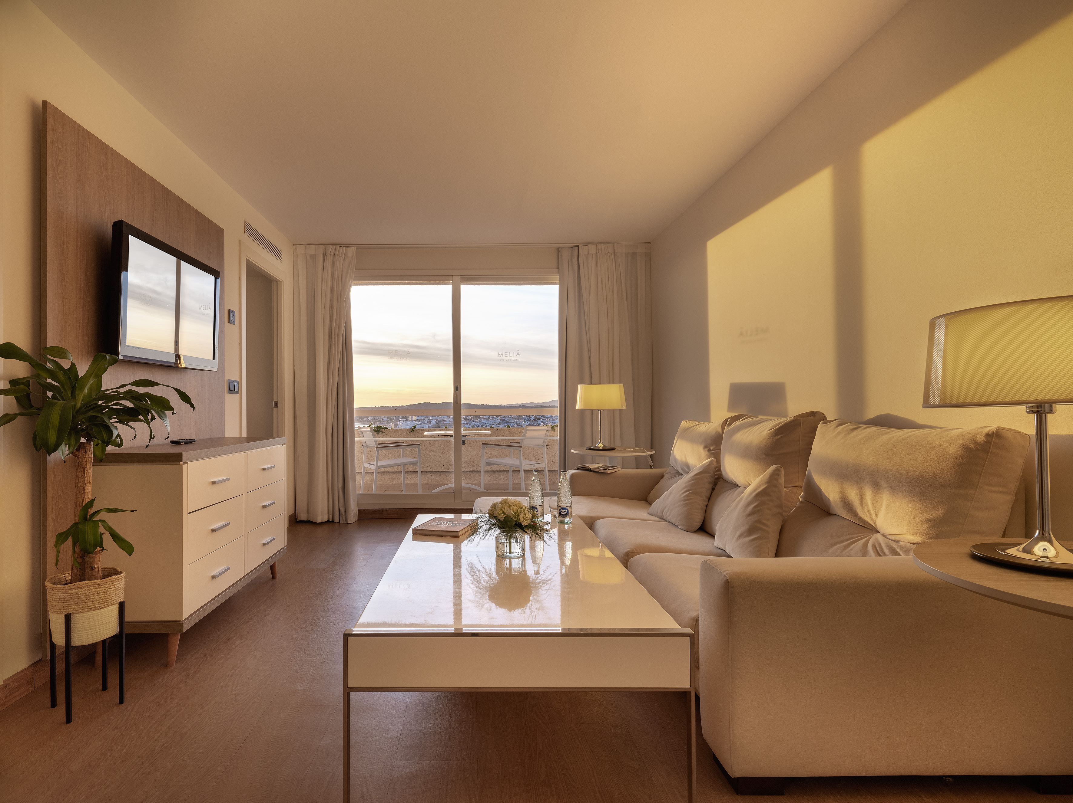 Modern hotel suite living room with sofa, TV, and balcony sunset view.