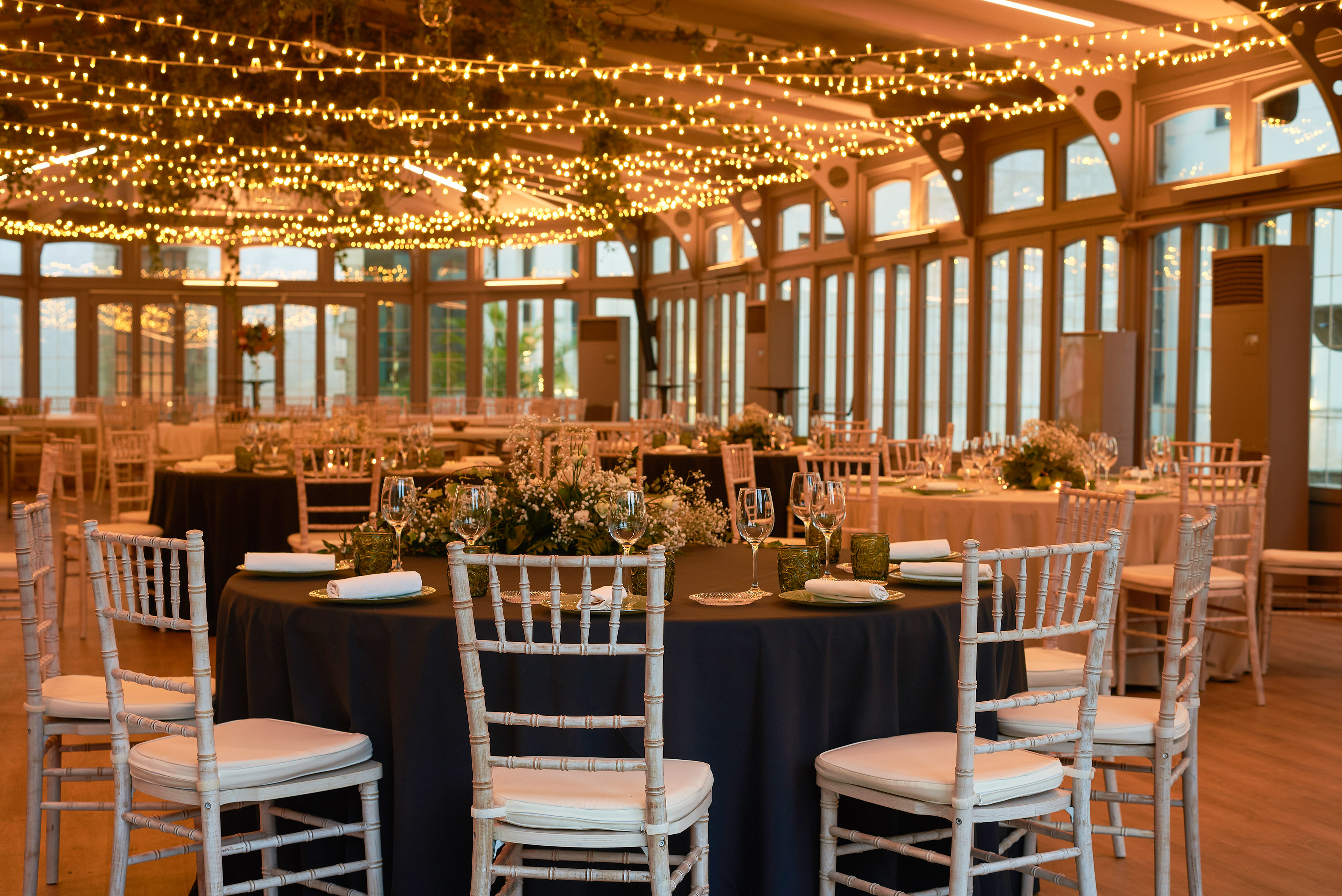 Elegant event hall with set dining tables, white chairs, and string lights.