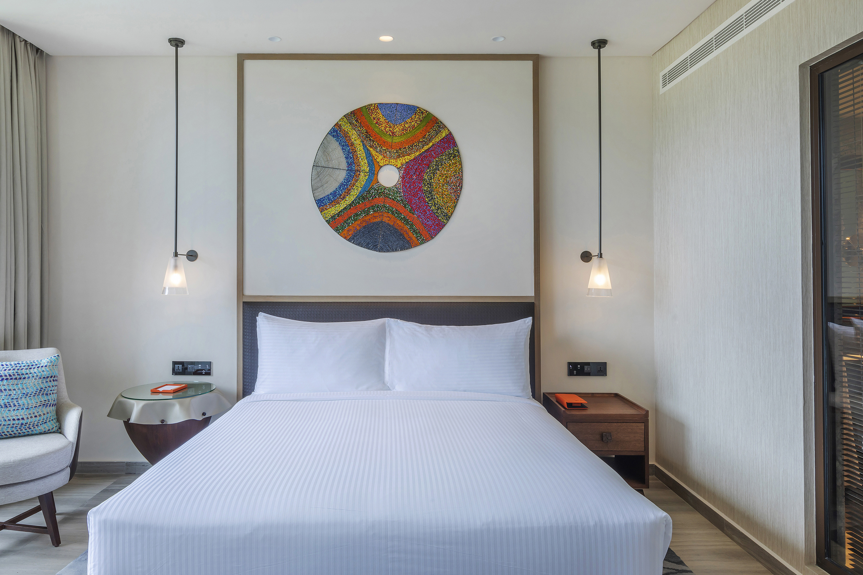 Modern hotel bedroom with white bed, colorful abstract art, and contemporary lighting.