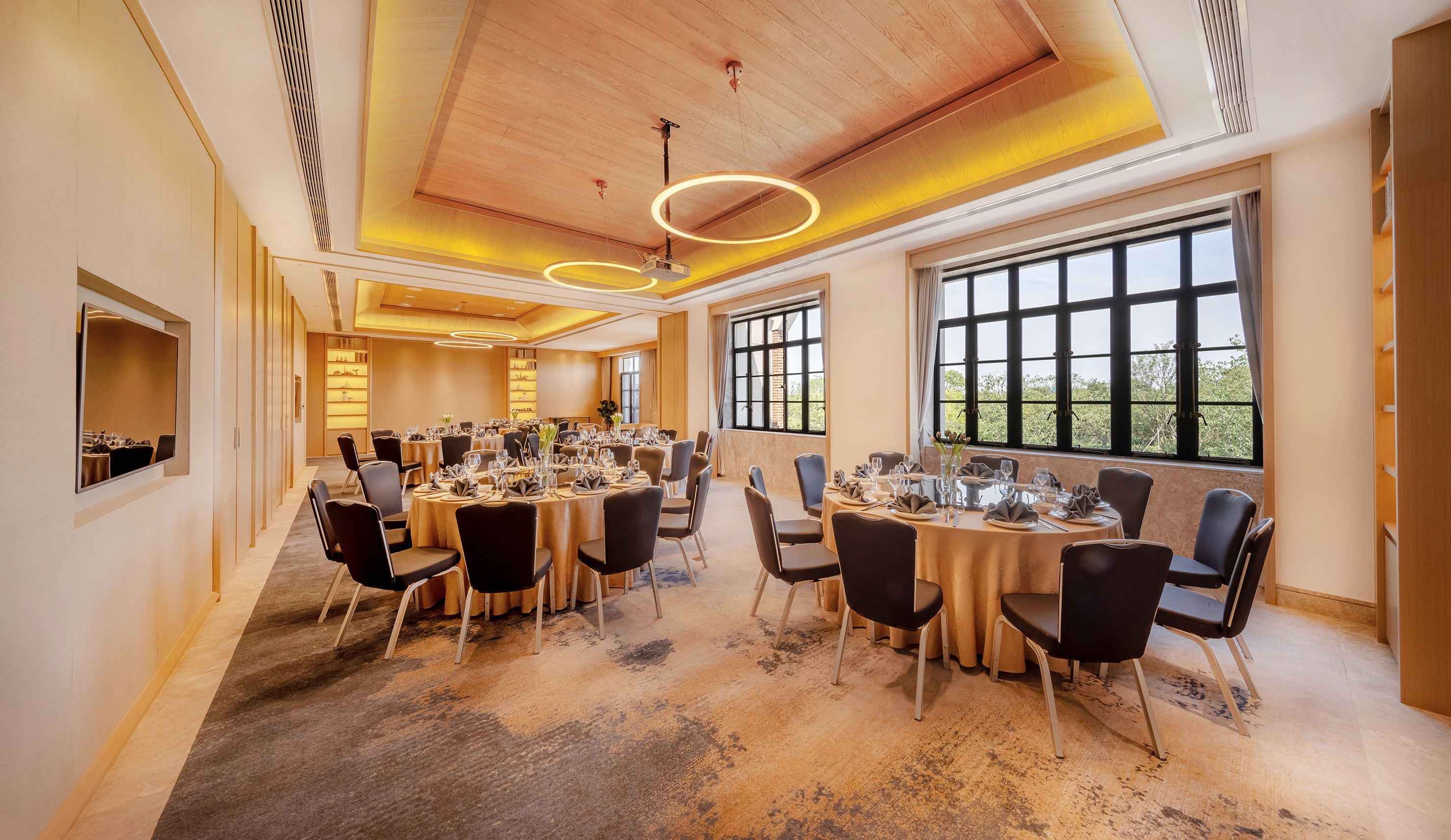 Modern banquet hall with set round tables, wood ceiling, and large scenic windows.