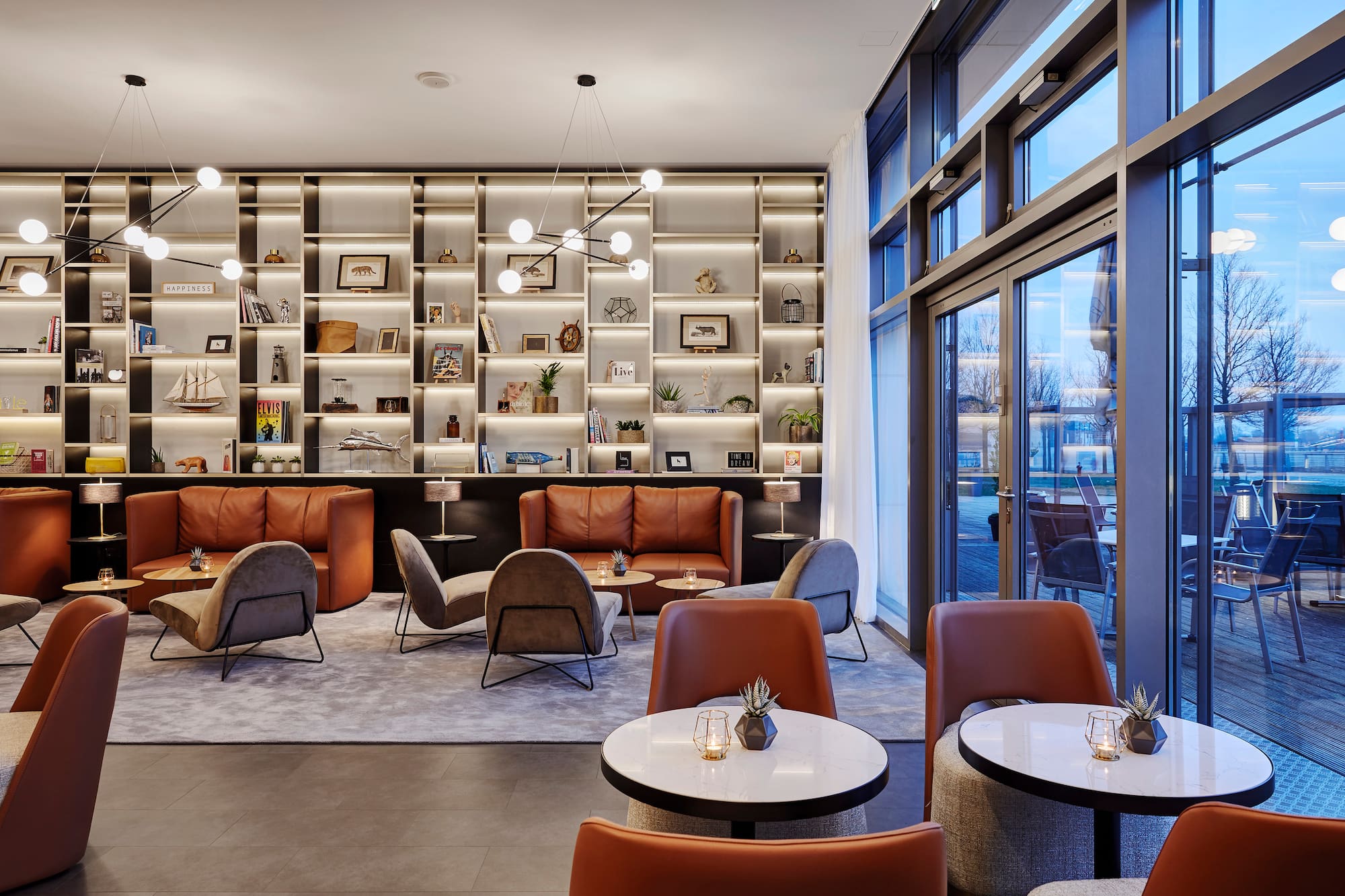 Modern lounge with brown leather sofas, illuminated shelves, and outdoor view.