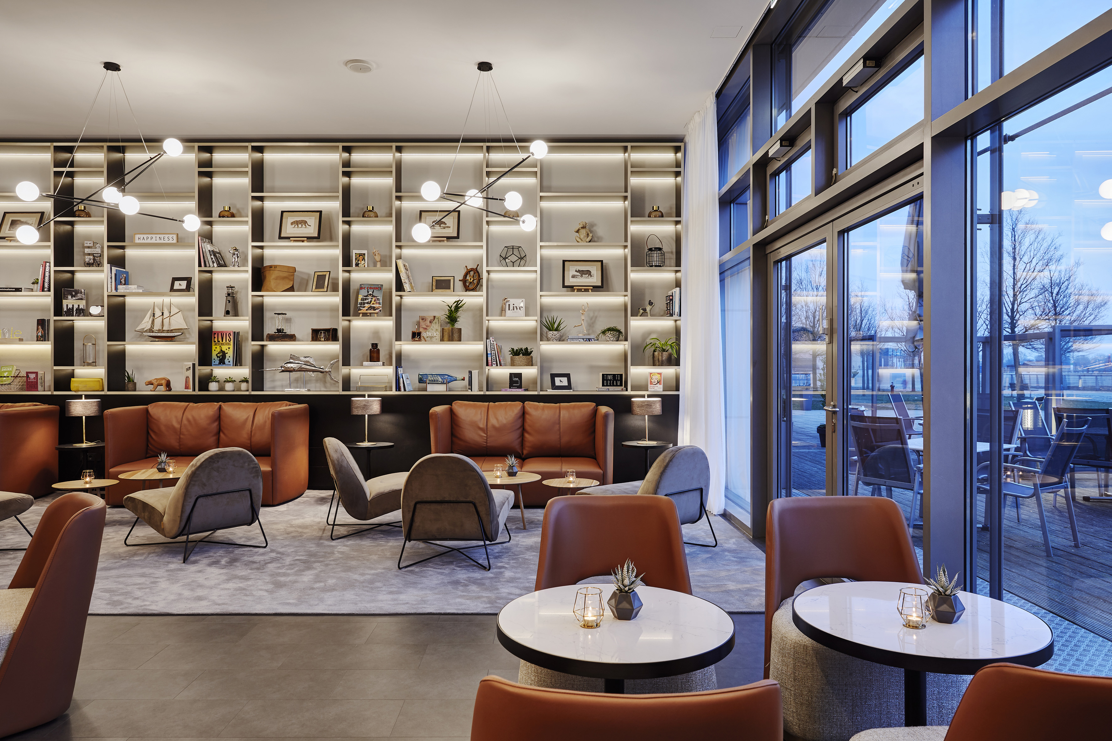 Modern lounge with brown leather sofas, illuminated shelves, and outdoor view.