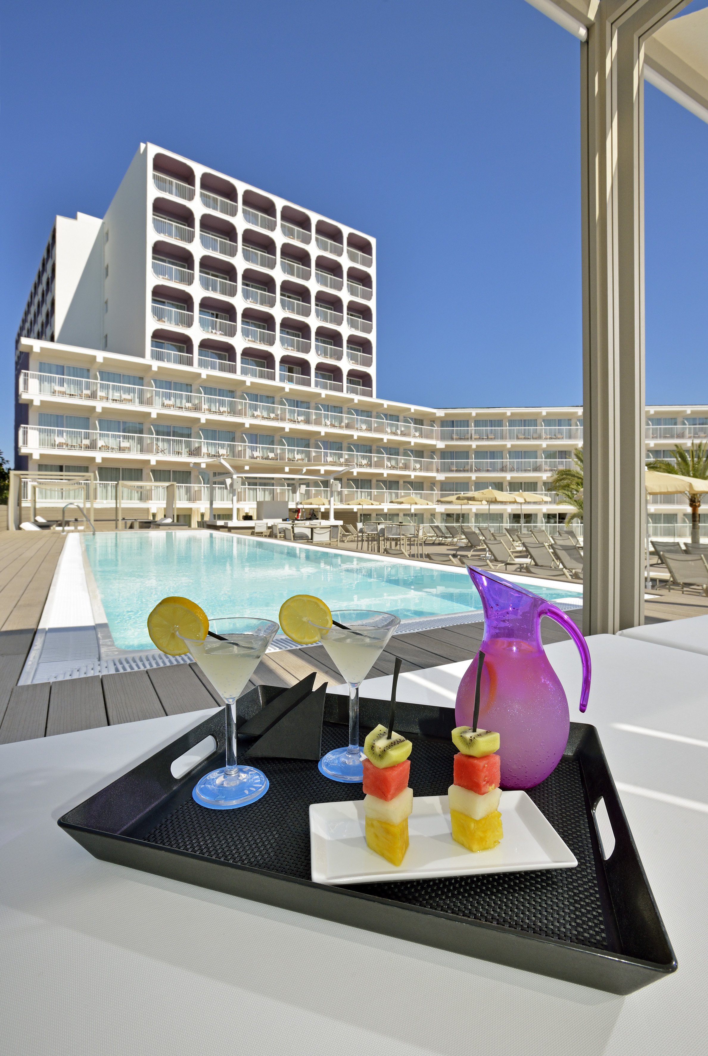 Poolside hotel with cocktails, fruit skewers, and a modern white building under blue sky.