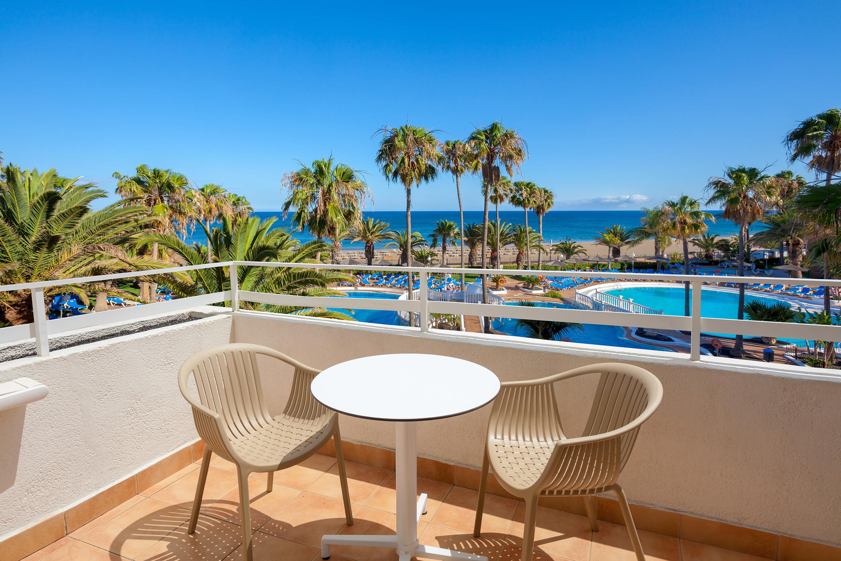 Balcony view of hotel pool, palm trees, beach, and ocean under blue sky.
