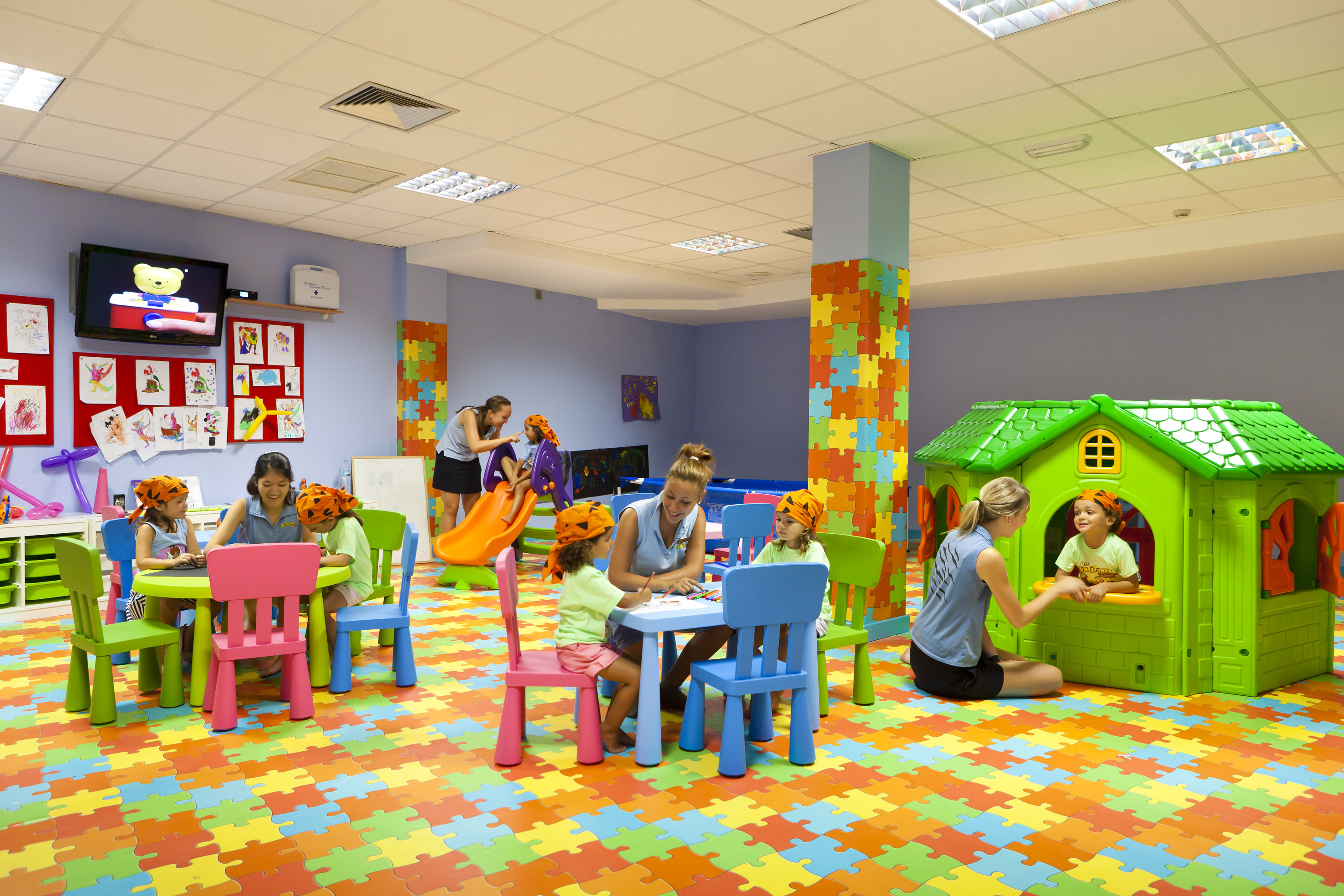 Colorful kids club playroom: children playing, drawing, and adults supervising.