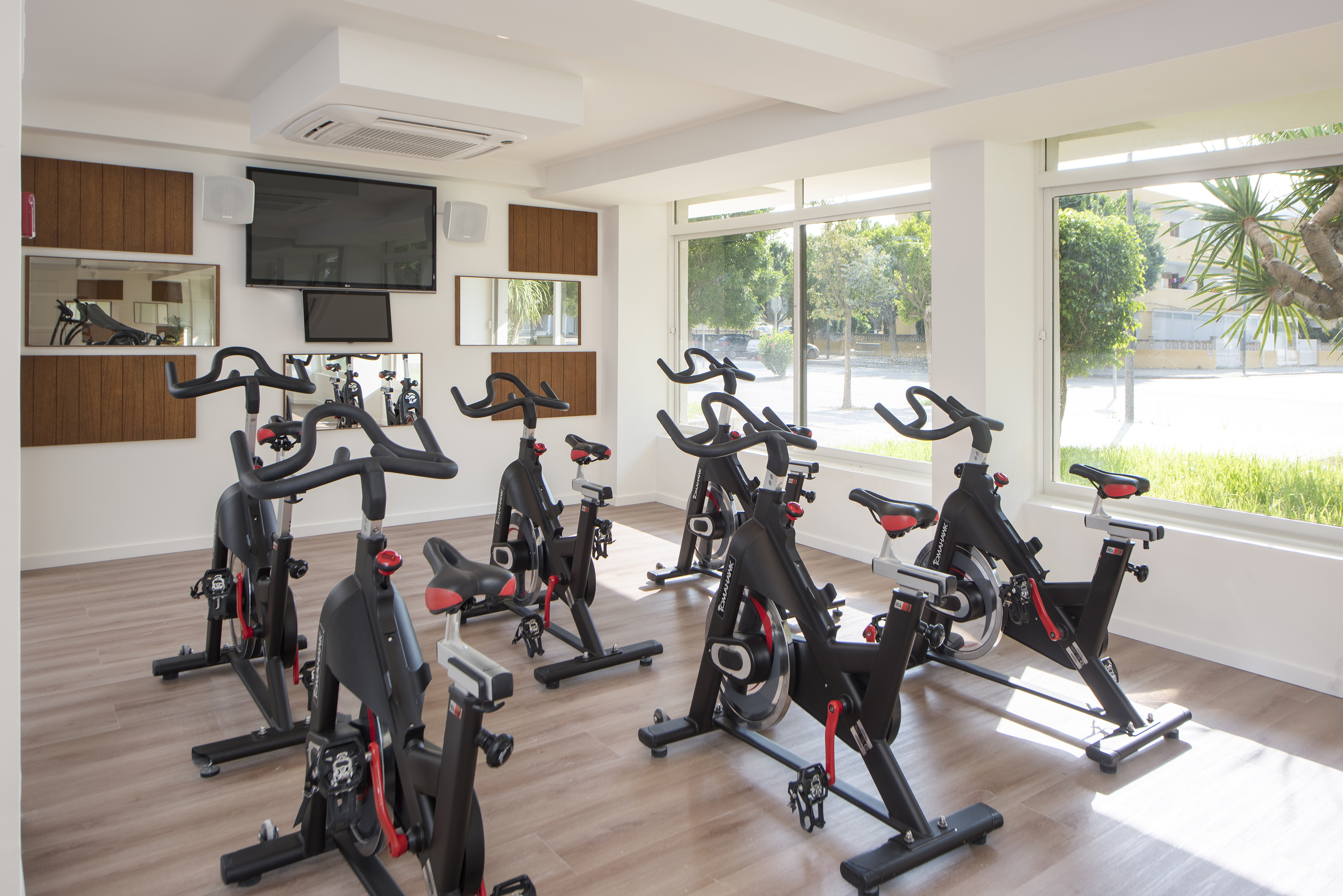 Bright spinning gym with black exercise bikes, wall TVs, and large windows.