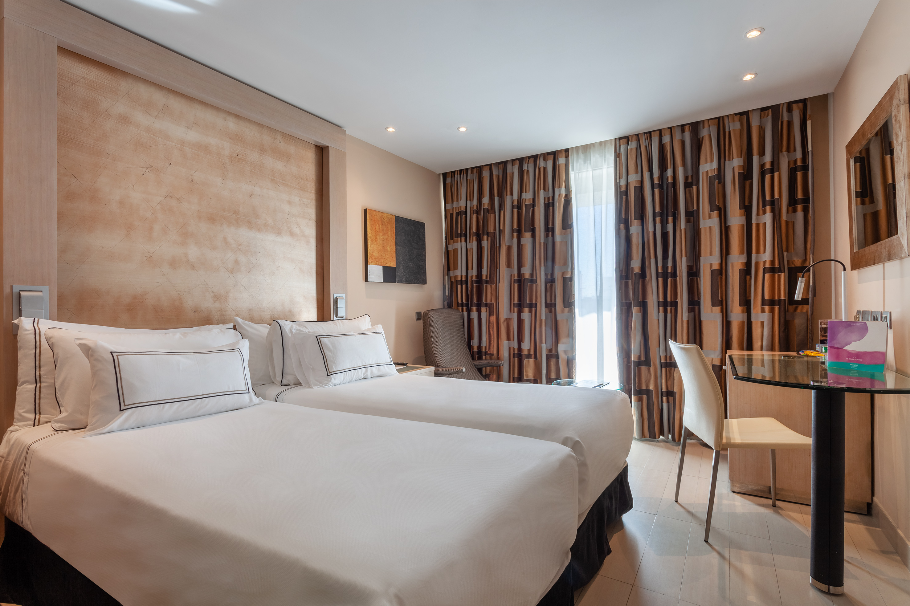 Modern hotel room with two twin beds, patterned curtains, and a wooden headboard.