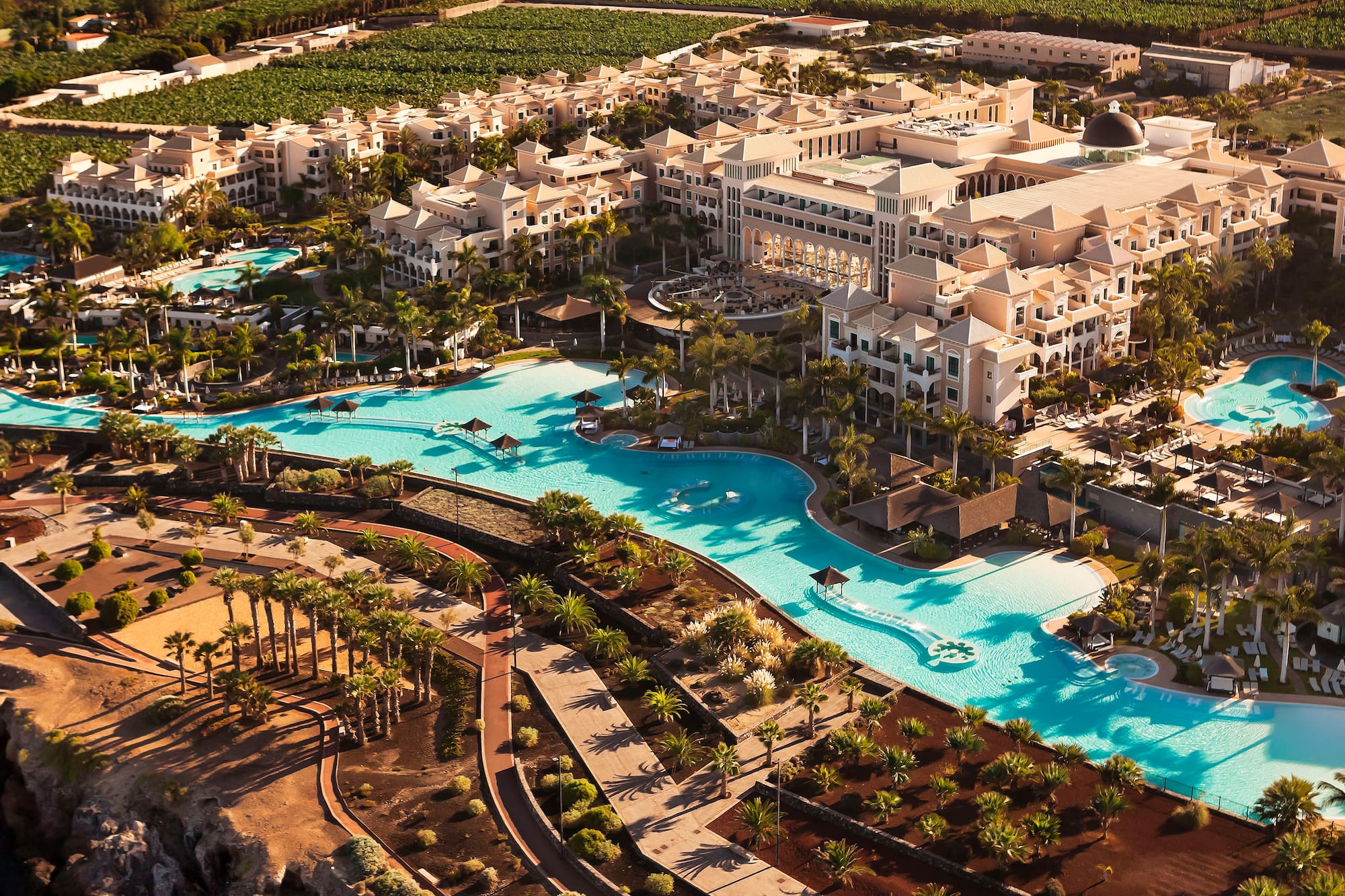 Aerial view: luxury hotel resort, winding pools, abundant palm trees, beige buildings.