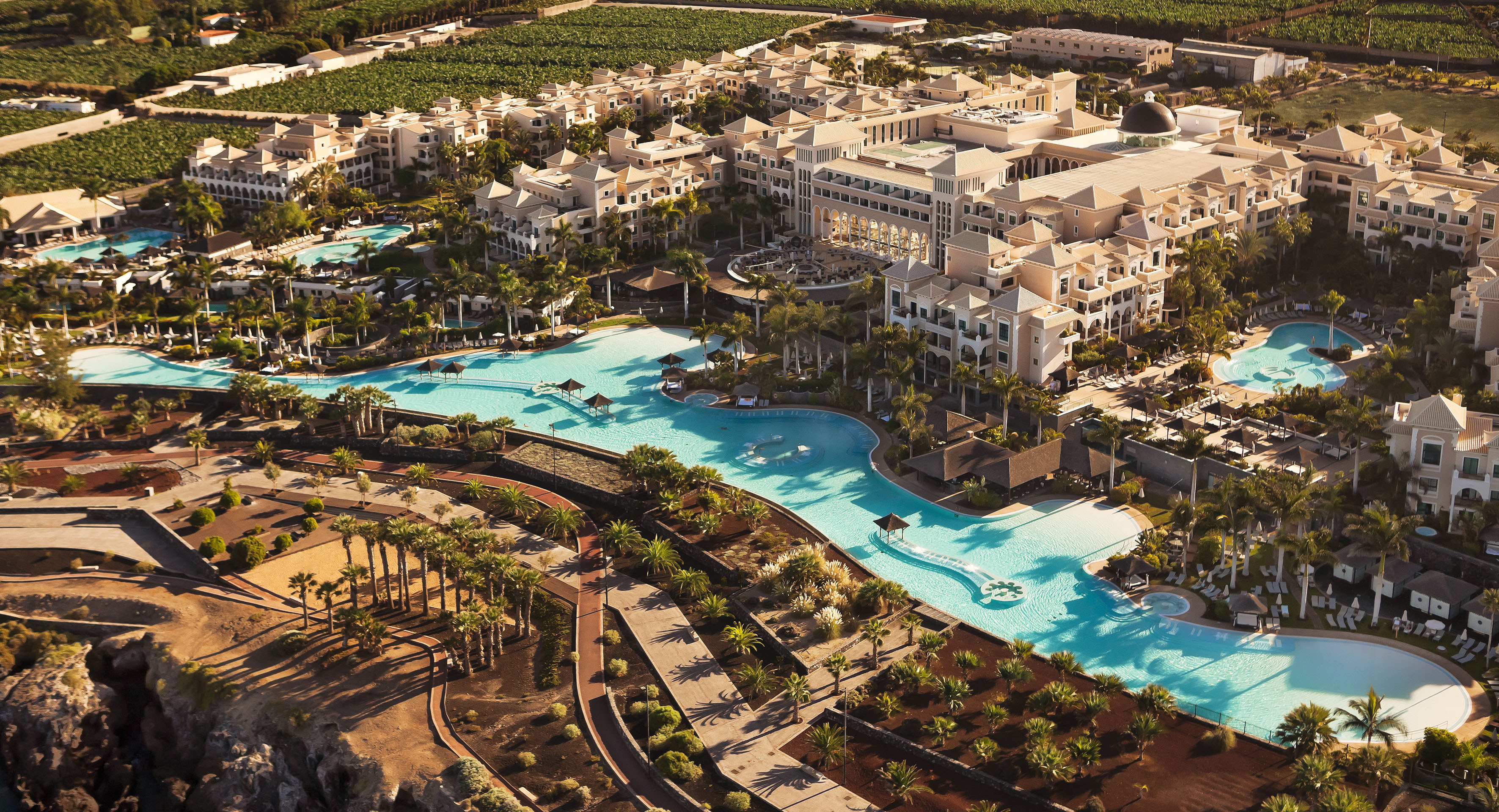 Aerial view: luxury hotel resort, winding pools, abundant palm trees, beige buildings.