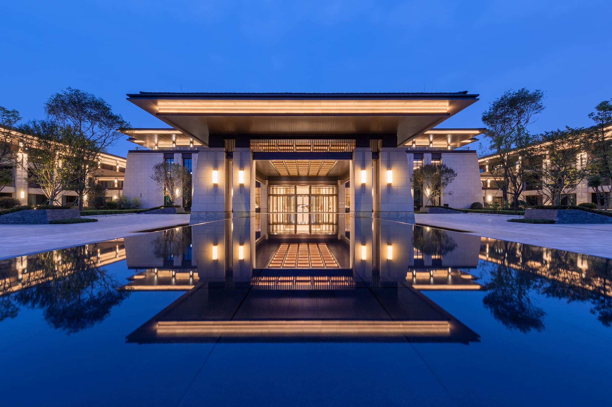 Illuminated modern luxury building entrance reflected in a tranquil pool at dusk.