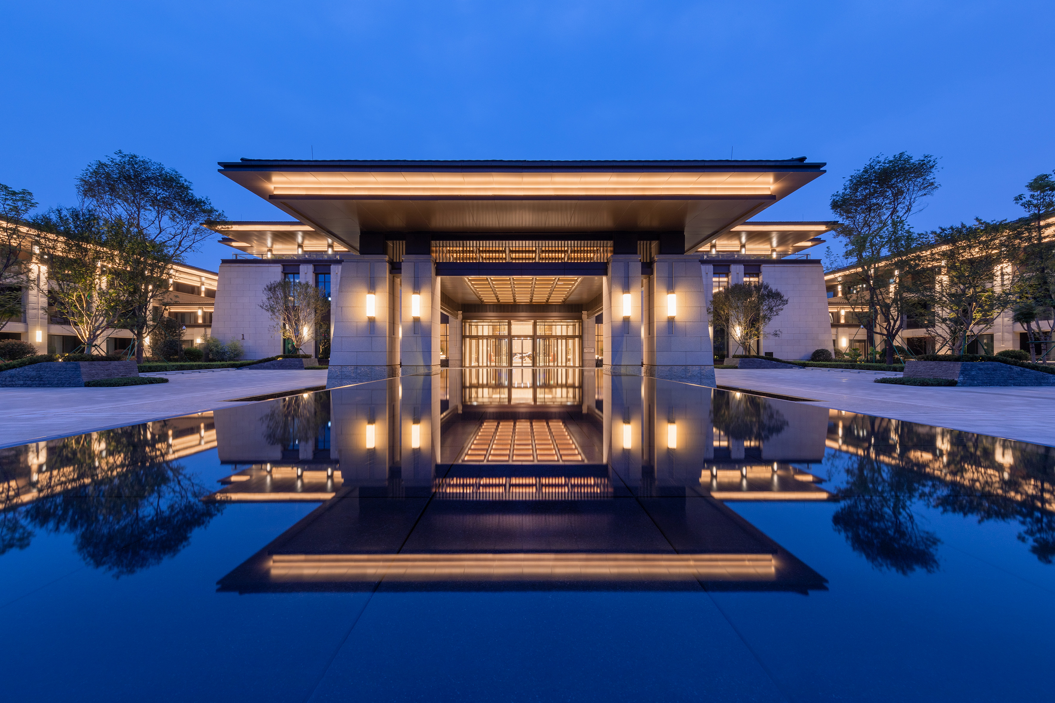 Illuminated modern luxury building entrance reflected in a tranquil pool at dusk.
