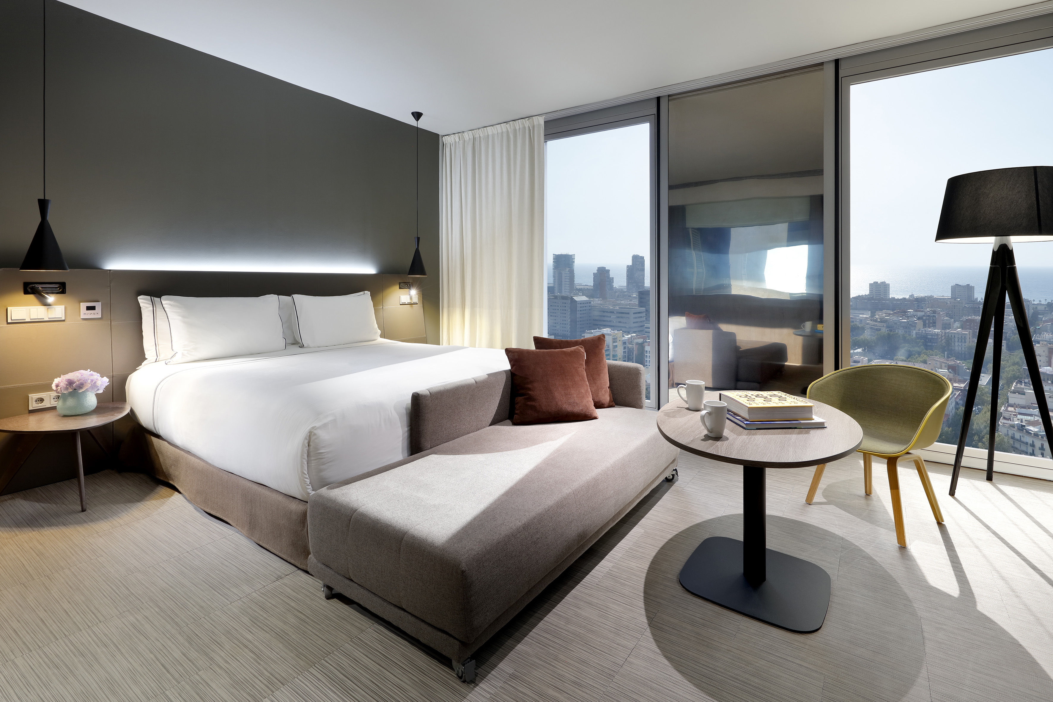 Modern hotel room with bed, city & ocean view from large window.