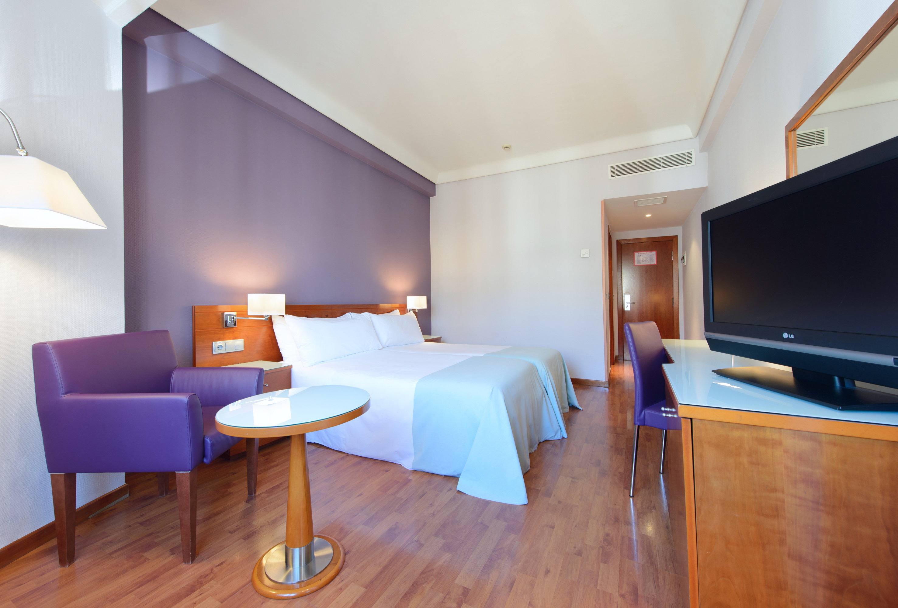 Modern hotel room with purple accent wall, two beds, TV, and wood floor.