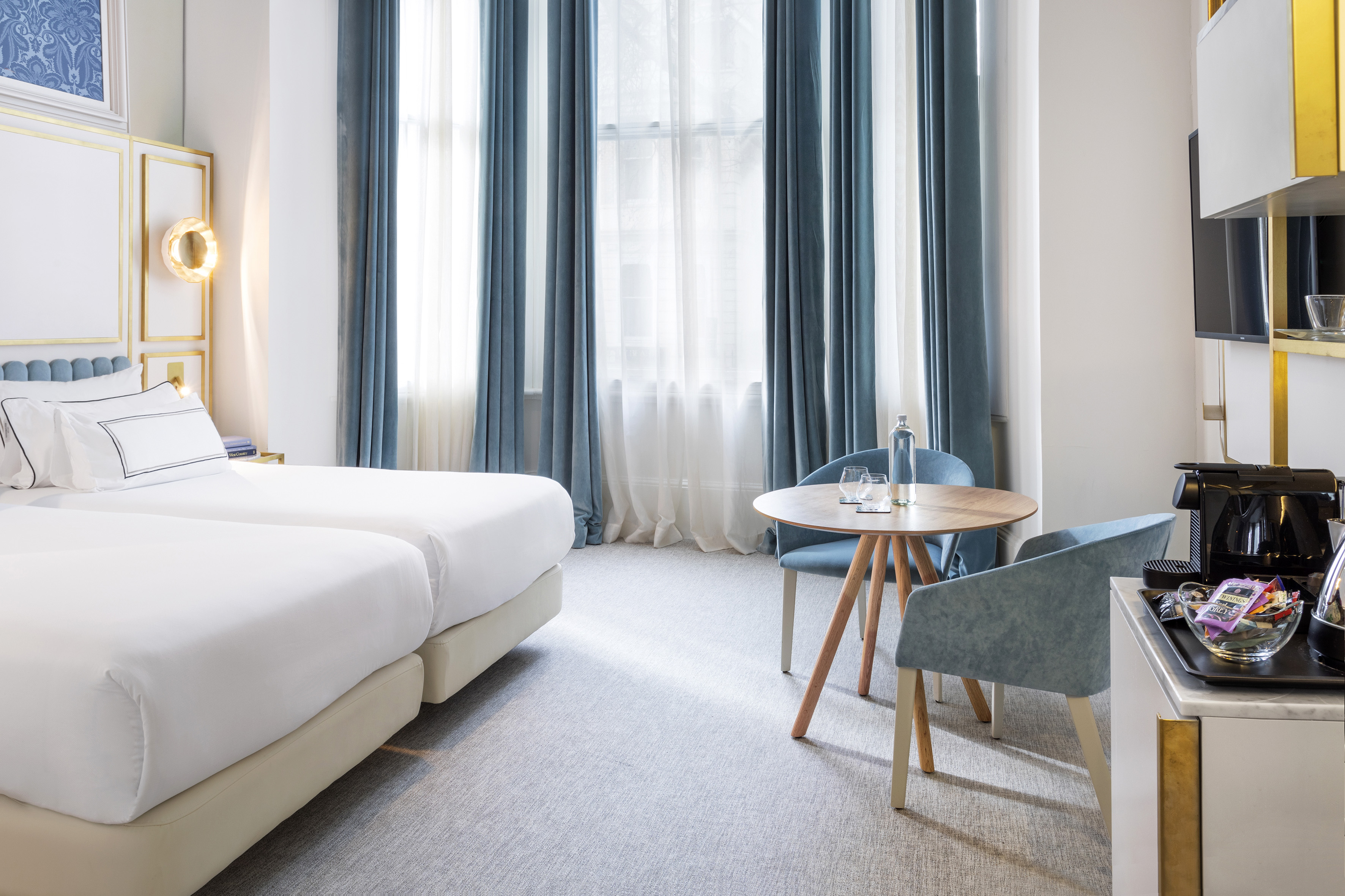 Modern hotel room with twin beds, blue curtains, and a coffee station.