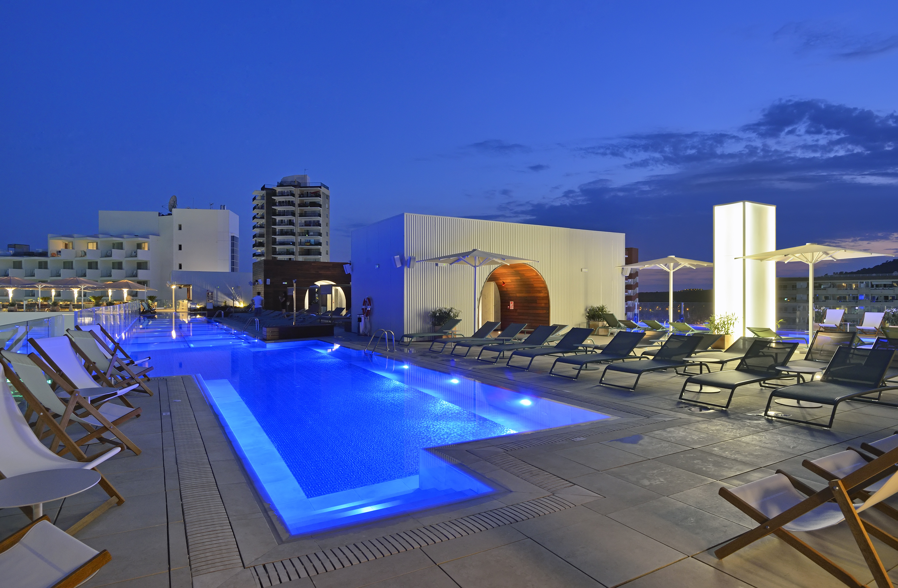 Blue lit rooftop pool at night with lounge chairs and hotel buildings.