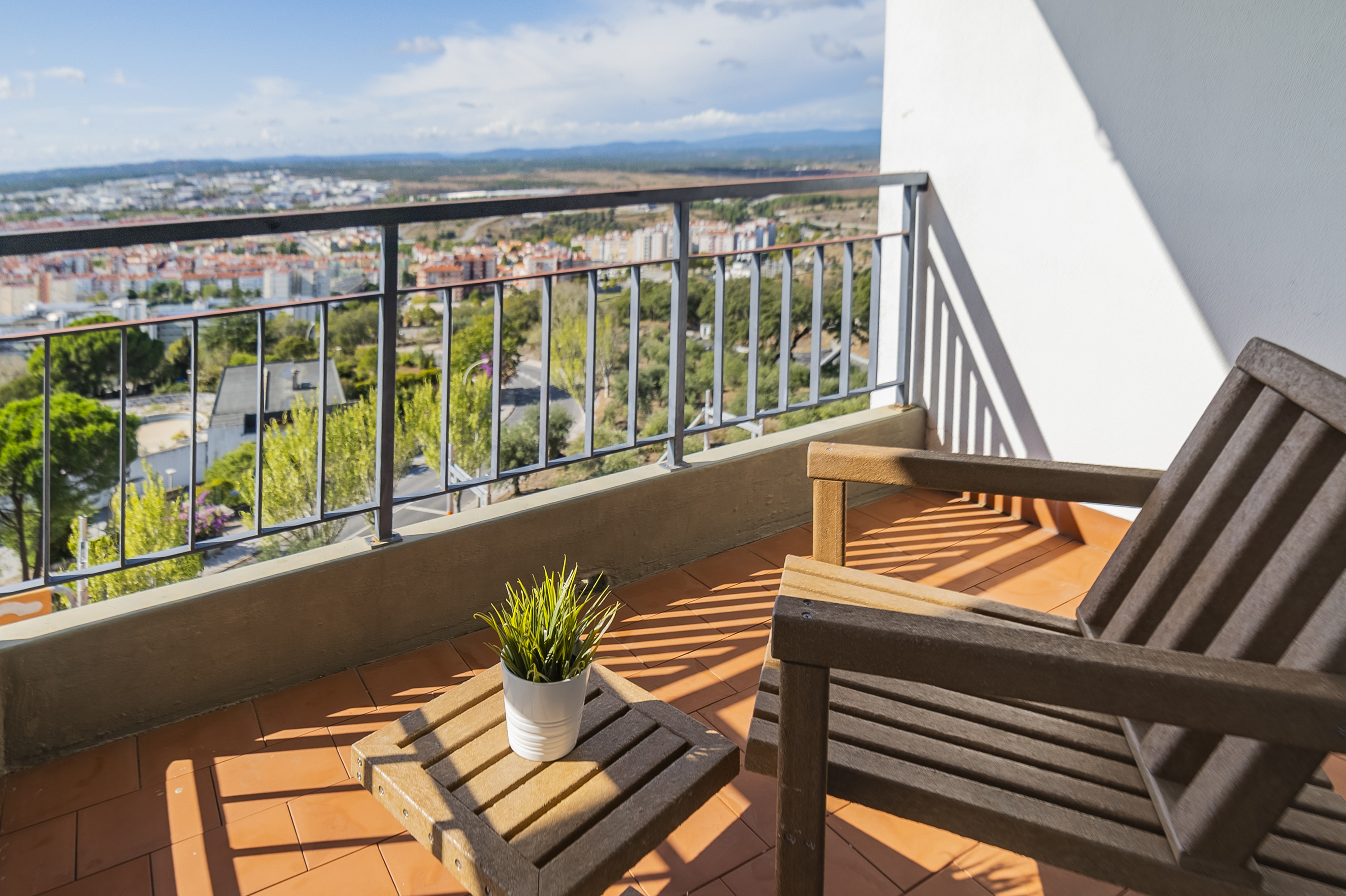 Sunny balcony with wooden furniture, potted plant, and expansive city view.