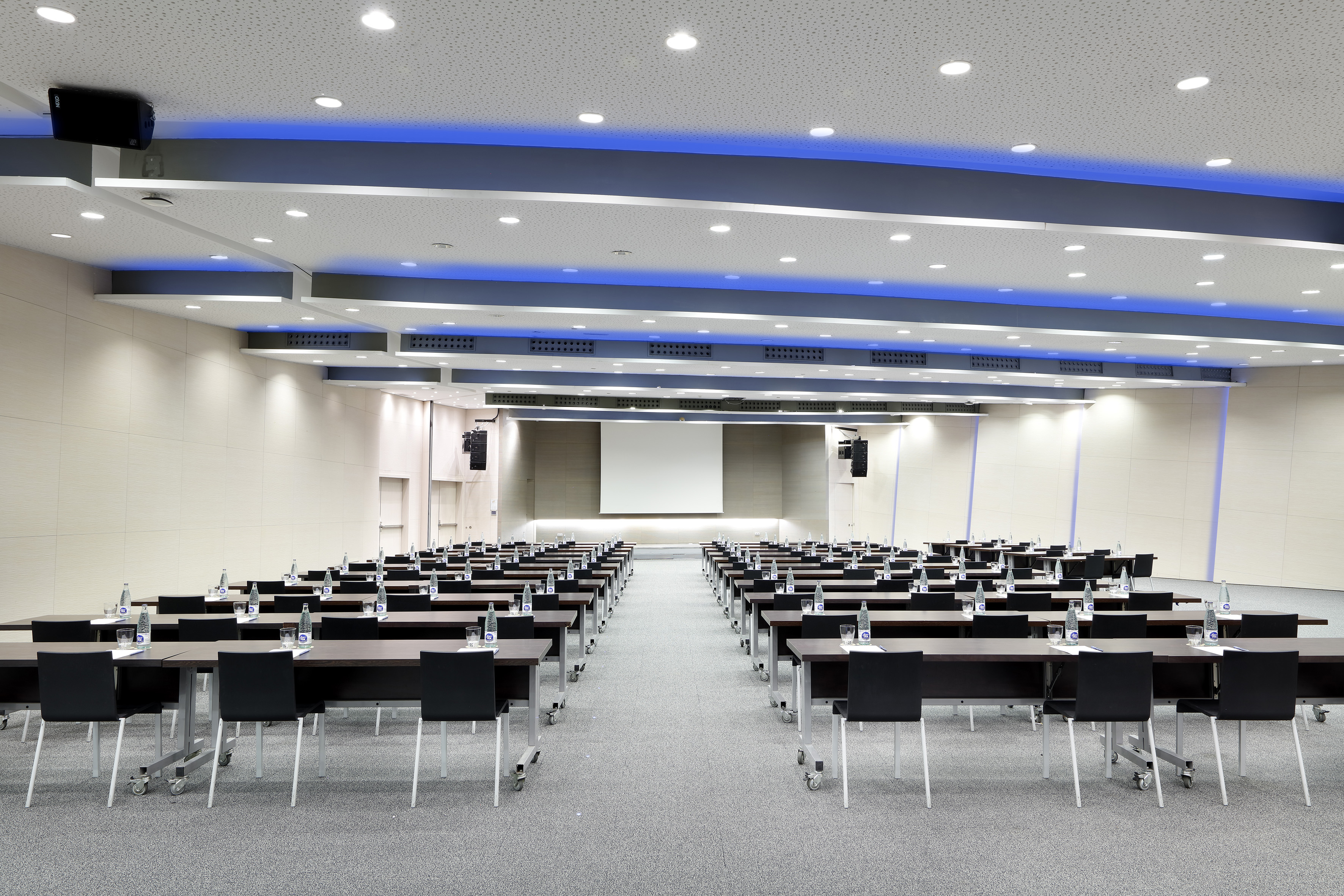 Modern conference room with rows of tables, chairs, projection screen, blue lights.