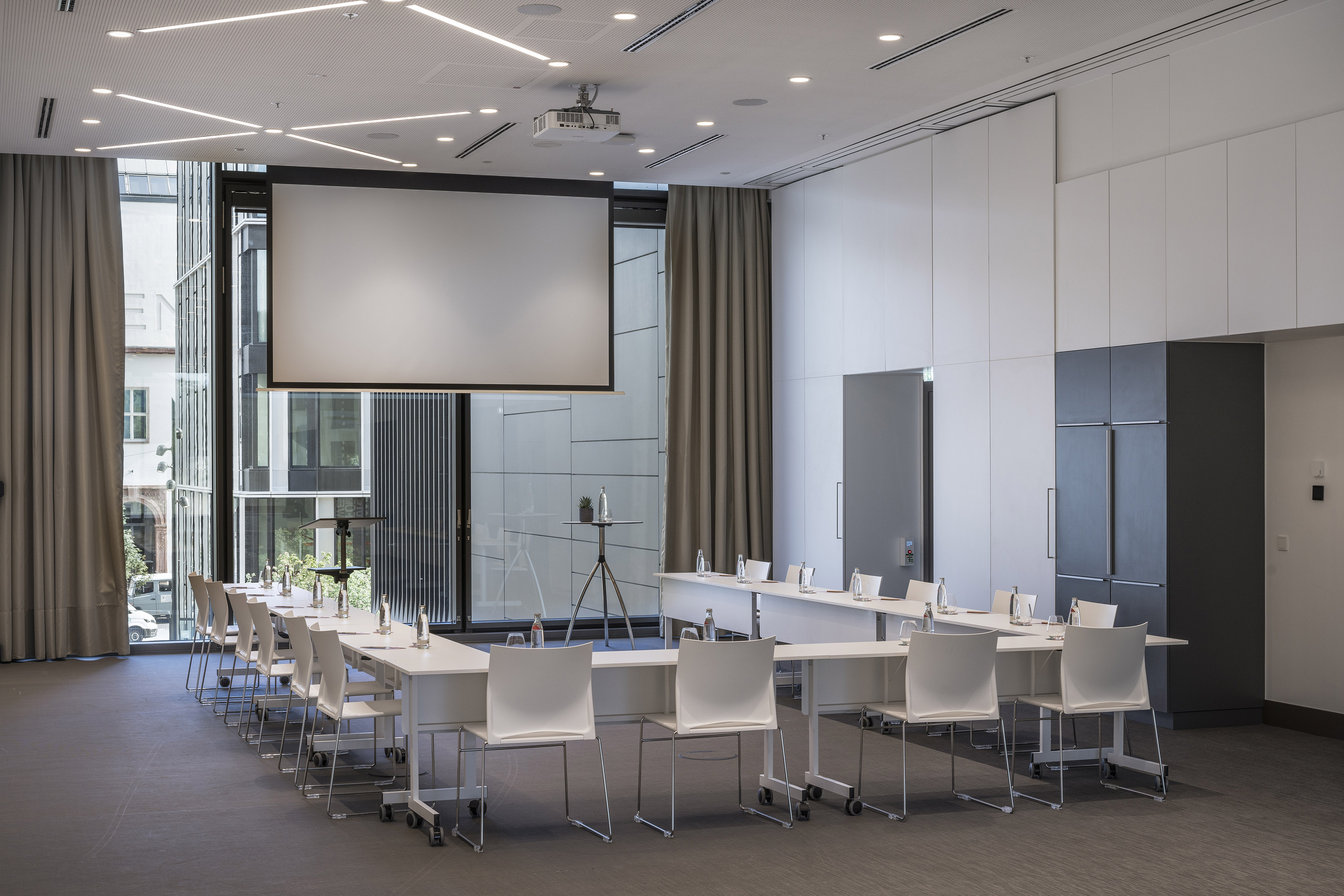 Modern U-shape meeting room with projector screen, white chairs, and windows.