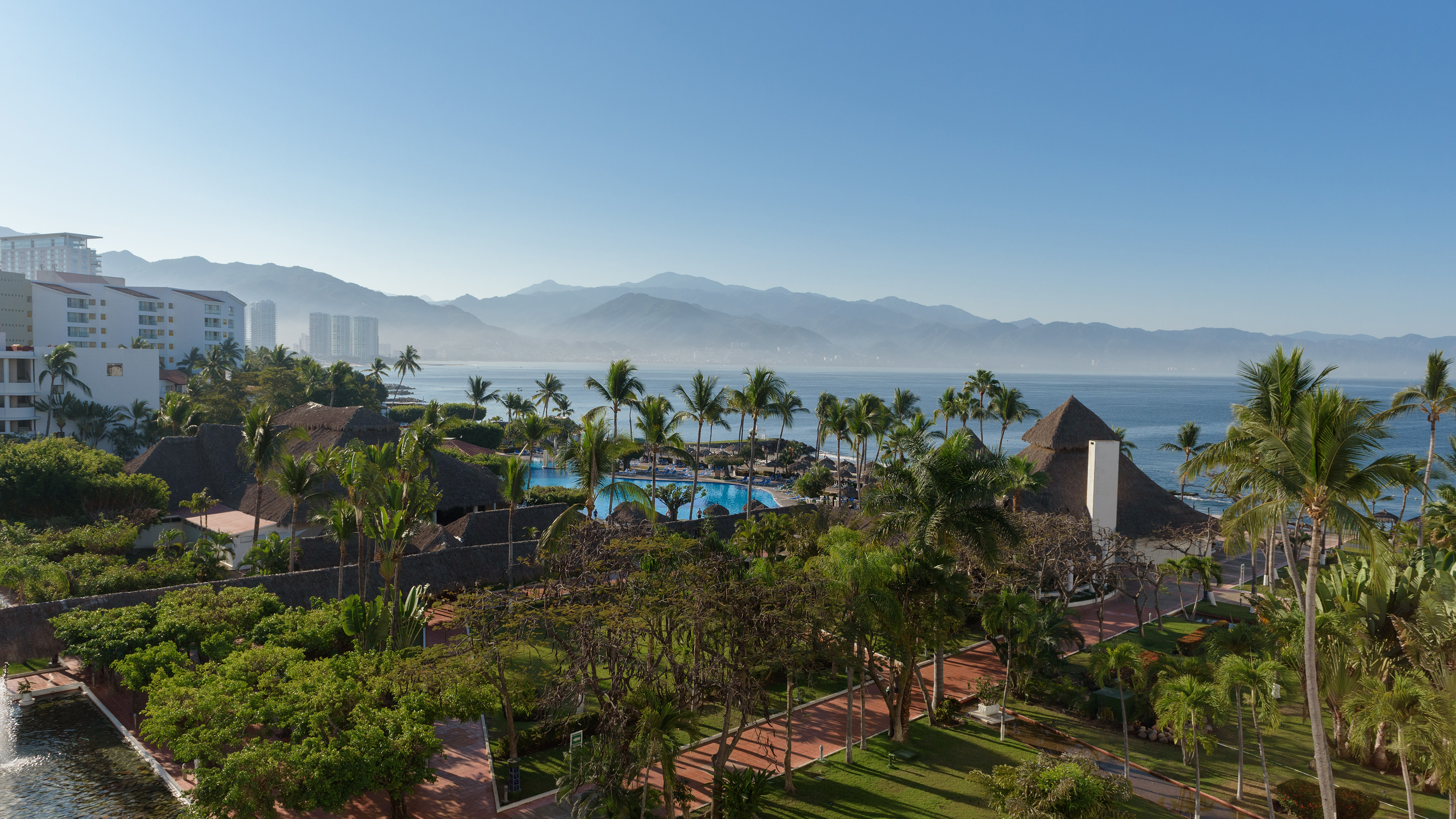 Tropical resort with ocean view, palm trees, swimming pools, and distant mountains.