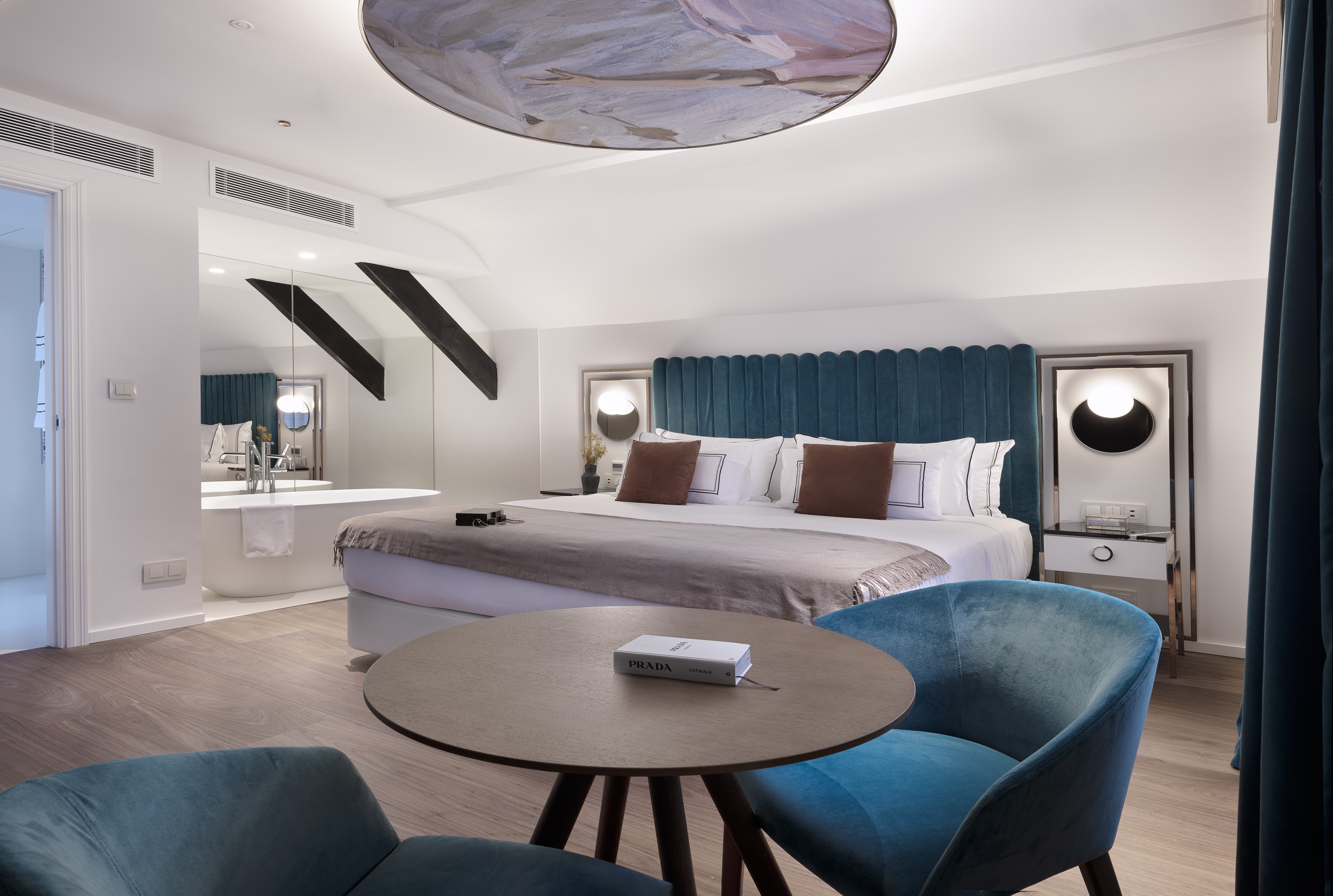 Modern luxury hotel room with teal velvet bed, round table, and freestanding tub.