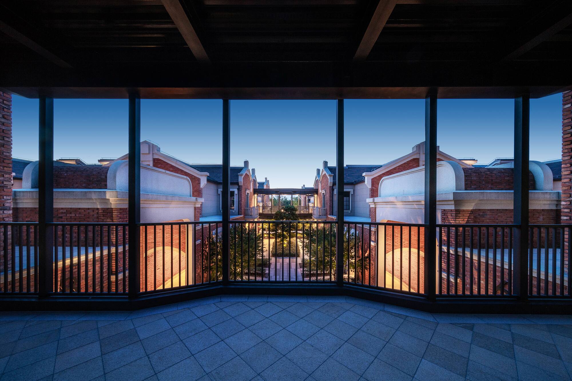 Symmetrical red brick buildings overlooking landscaped courtyard from balcony at dusk.