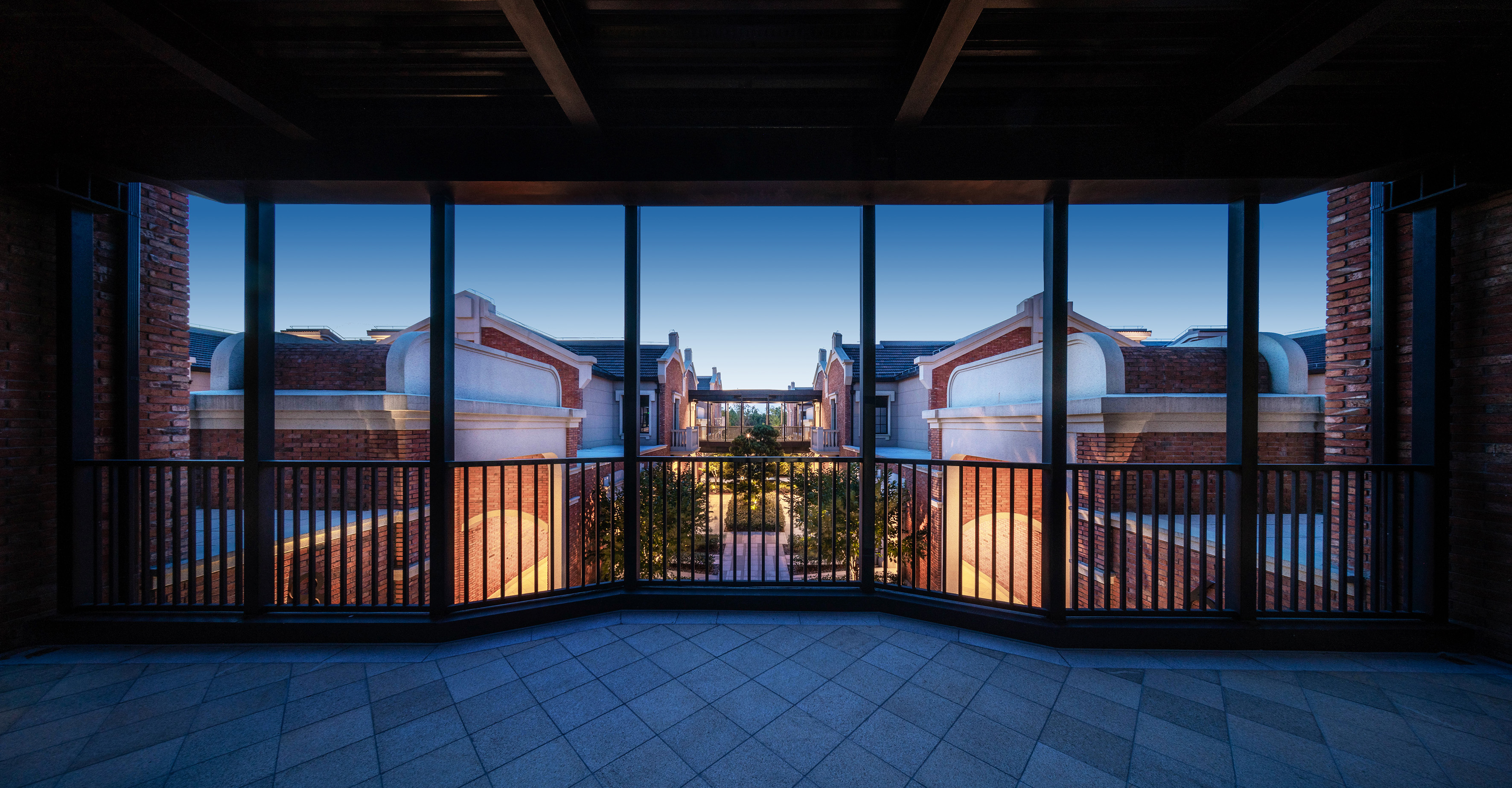Symmetrical red brick buildings overlooking landscaped courtyard from balcony at dusk.