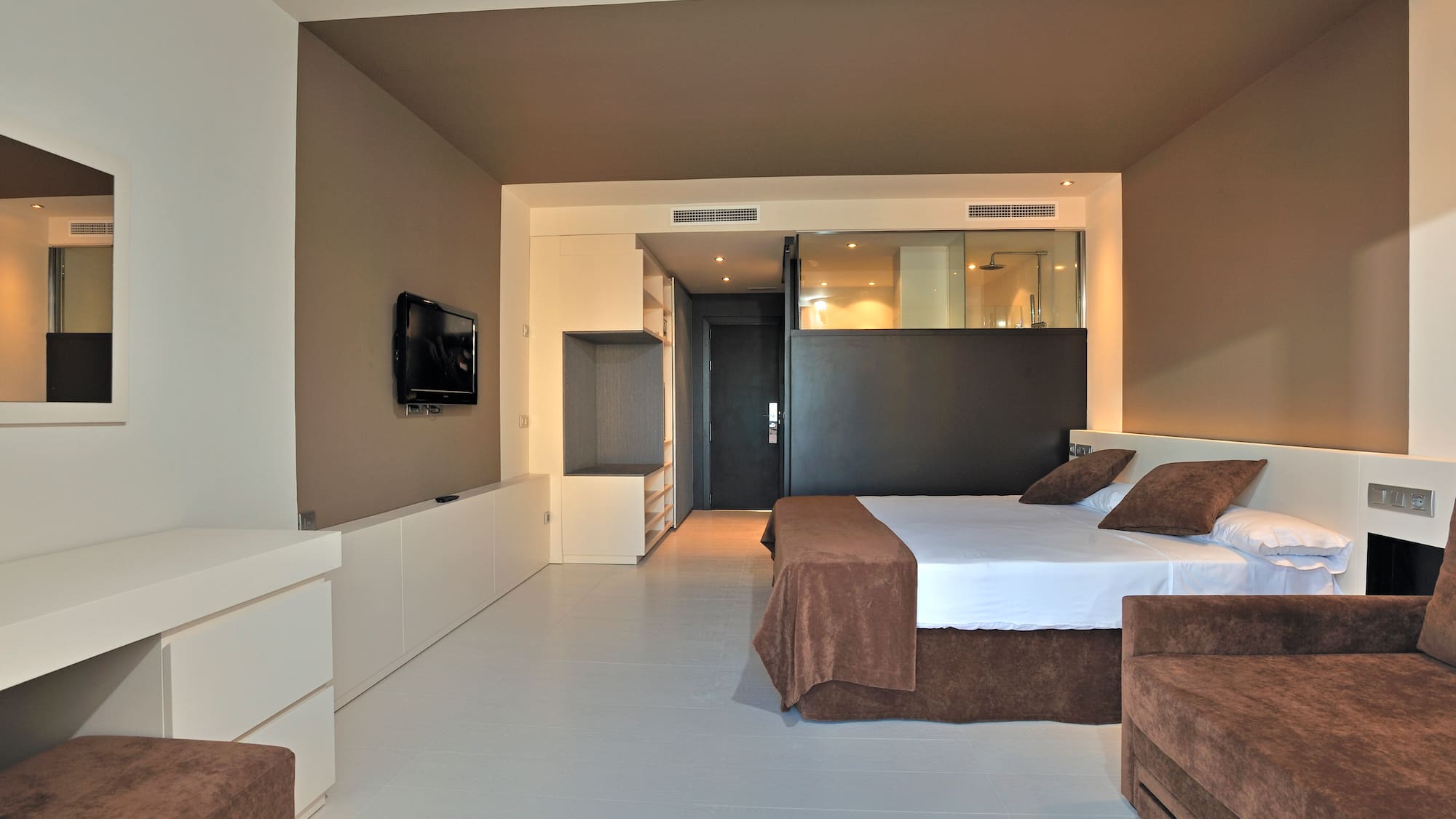 Contemporary hotel room with brown king bed, sofa, TV, and glass bathroom.