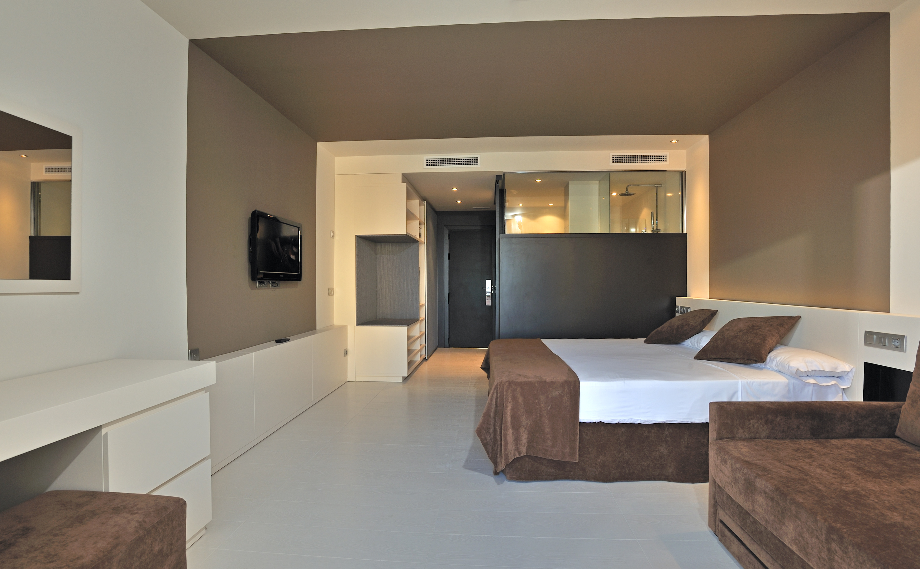 Contemporary hotel room with brown king bed, sofa, TV, and glass bathroom.